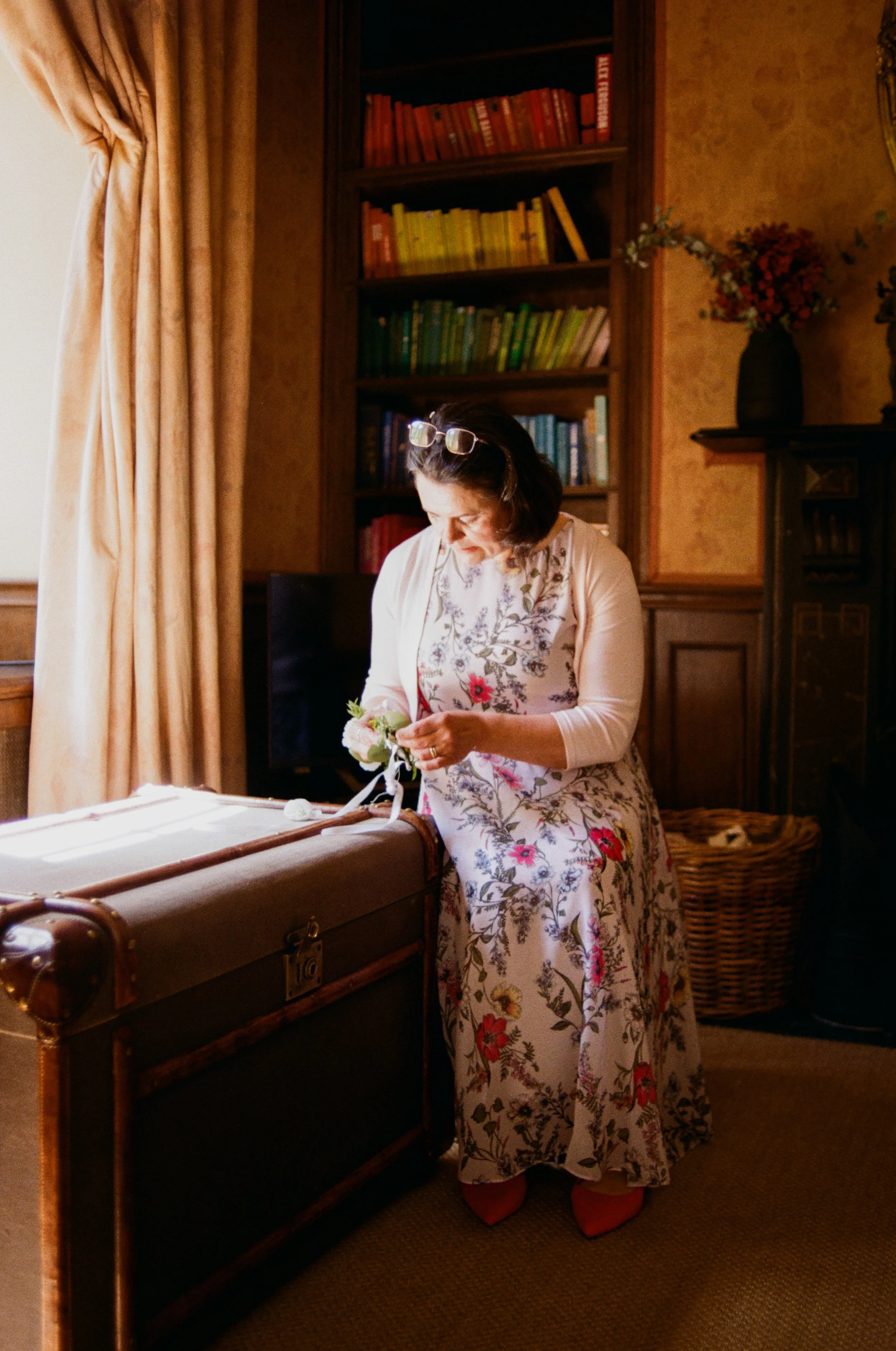 A woman dressed in a floral dress and red shoes, standing by a vintage trunk in a cozy, traditional room with a bookshelf and a vase of flowers.