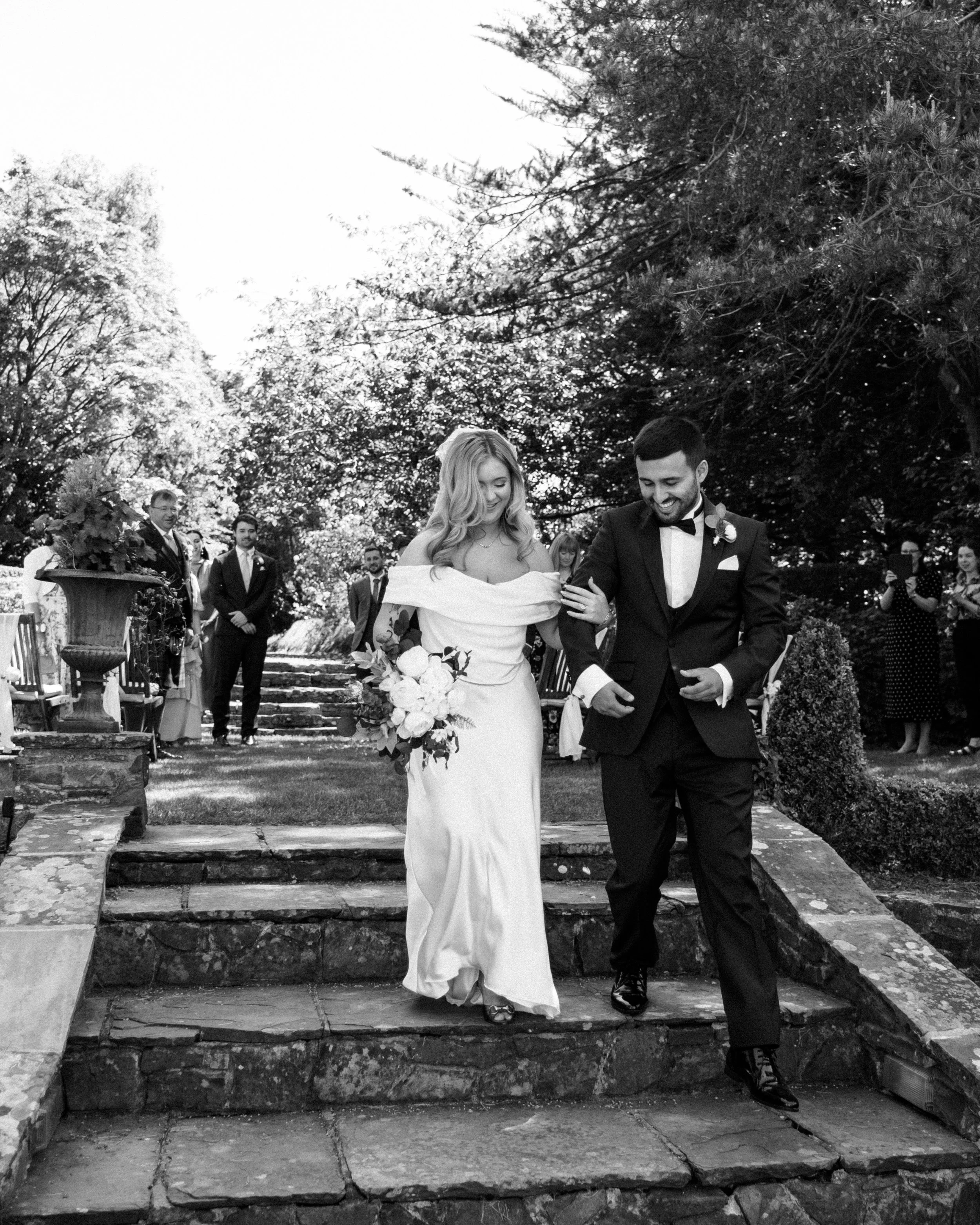 A wedding ceremony outdoors with a bride and groom walking down stone steps, surrounded by trees and guests in formal attire.