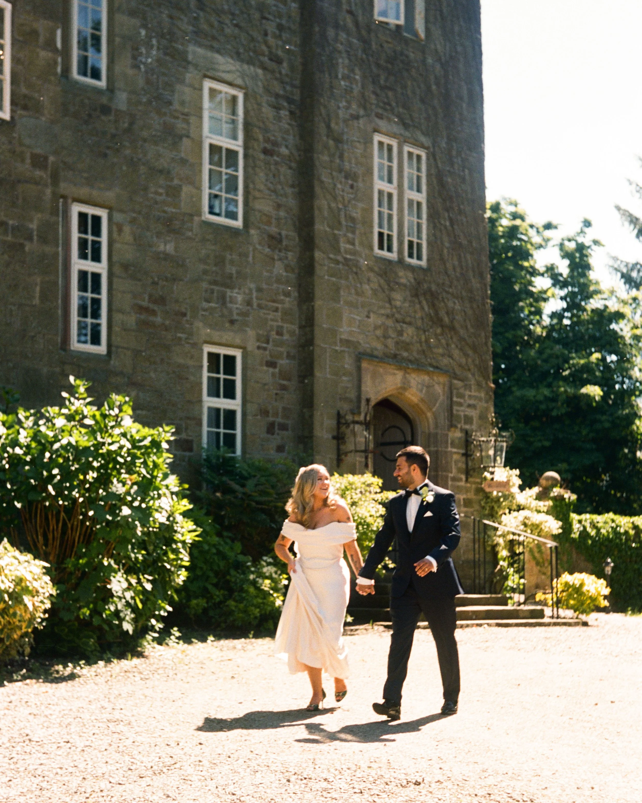 A bride and groom holding hands and walking outdoors on a bright, sunny day. The bride is in a white off-shoulder gown and the groom in a black tuxedo. They are near a stone building with multiple windows and surrounded by lush green bushes and trees