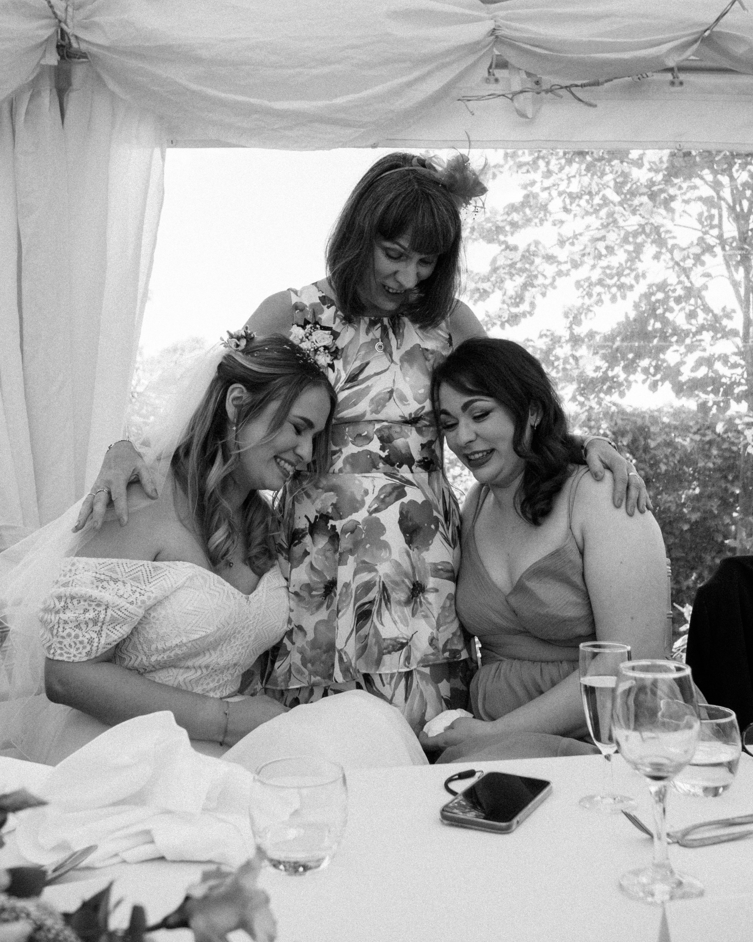 Four women are sitting together at a table outdoors under a canopy, smiling and hugging each other. The table has empty glasses, a smartphone, and scattered napkins. The background shows trees and natural light.
