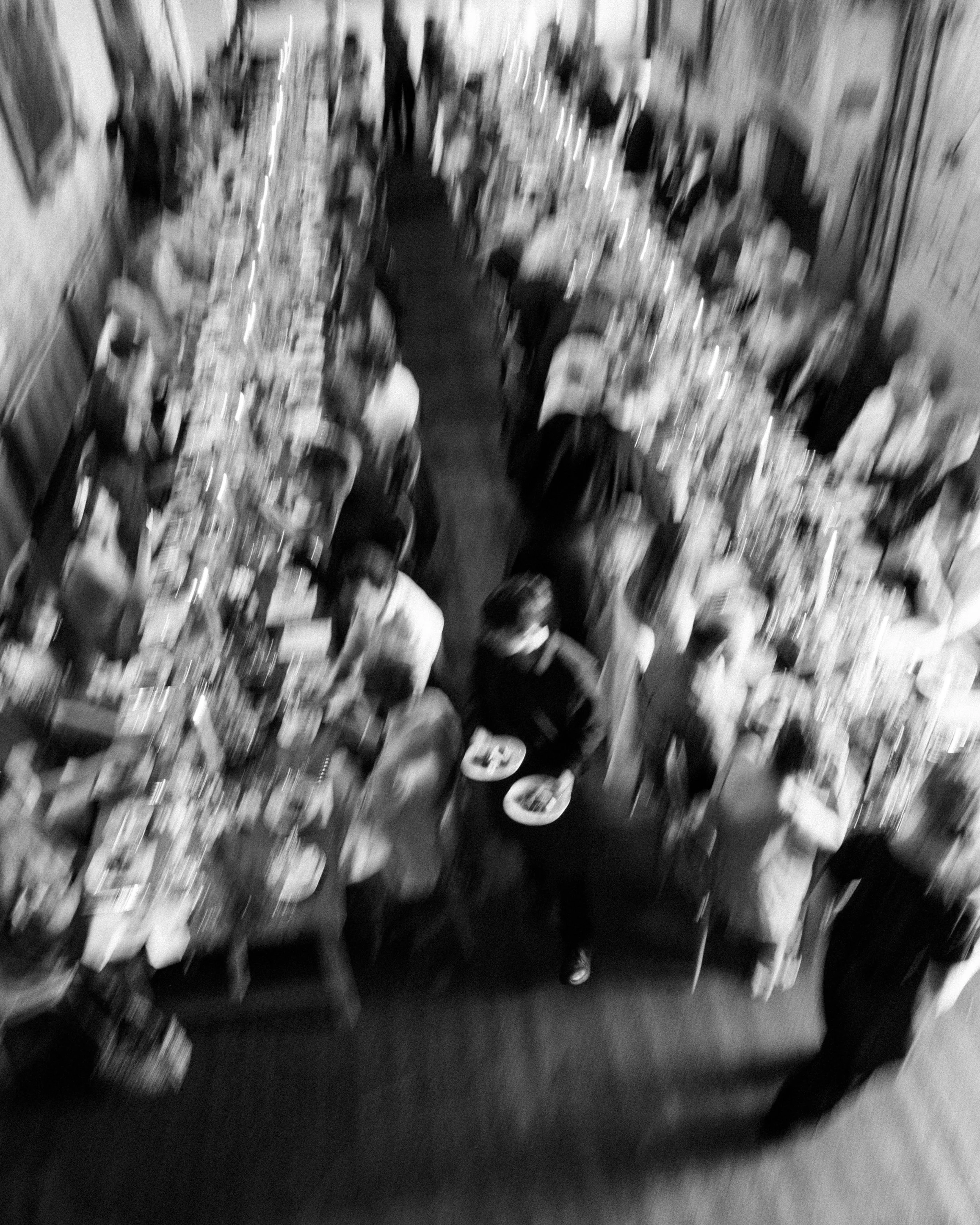 Blurred image of people walking through a hallway lined with shelves of books or magazines.