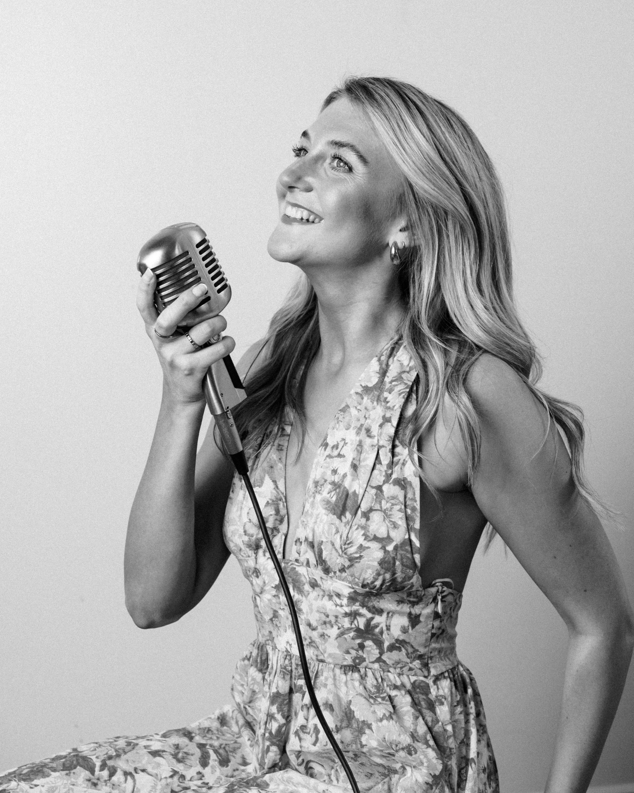A woman with long wavy hair, wearing a patterned sleeveless dress, smiling and holding a retro-style microphone, appearing to sing or speak.