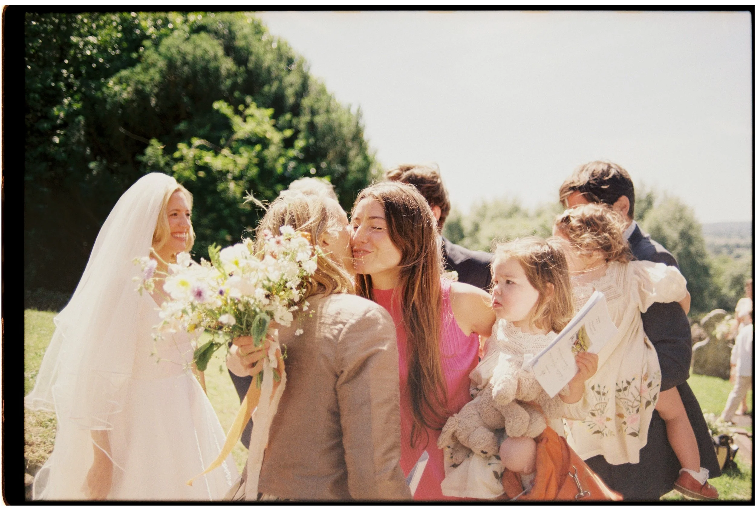 A group of people, including a bride with a veil and bouquet, gathering outdoors on a sunny day, smiling and greeting each other.