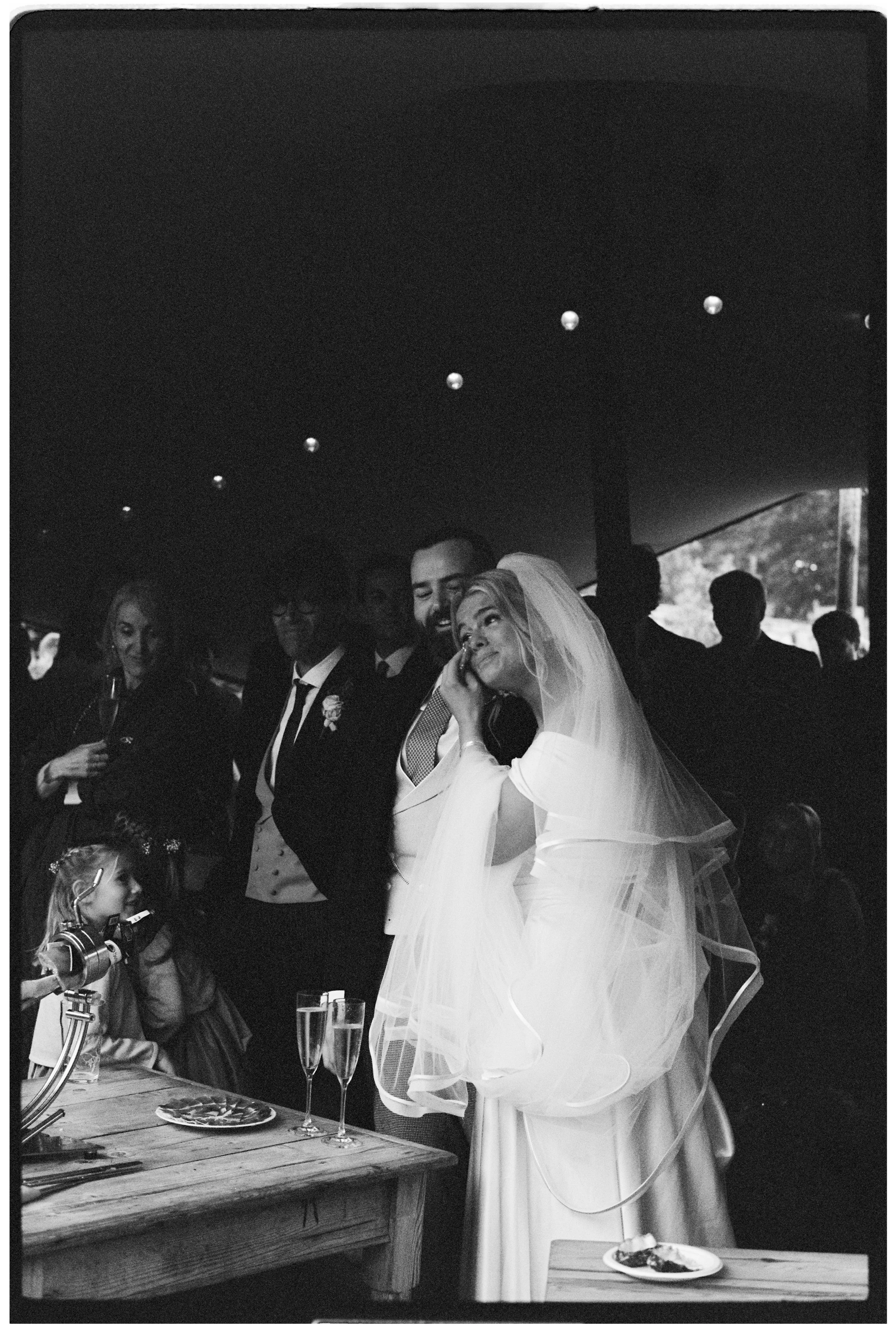 A wedding reception with a bride and groom smiling and celebrating among guests. The bride is wearing a wedding dress and veil, and the groom is in a tuxedo. There are champagne glasses, a cake, and a decorated table.