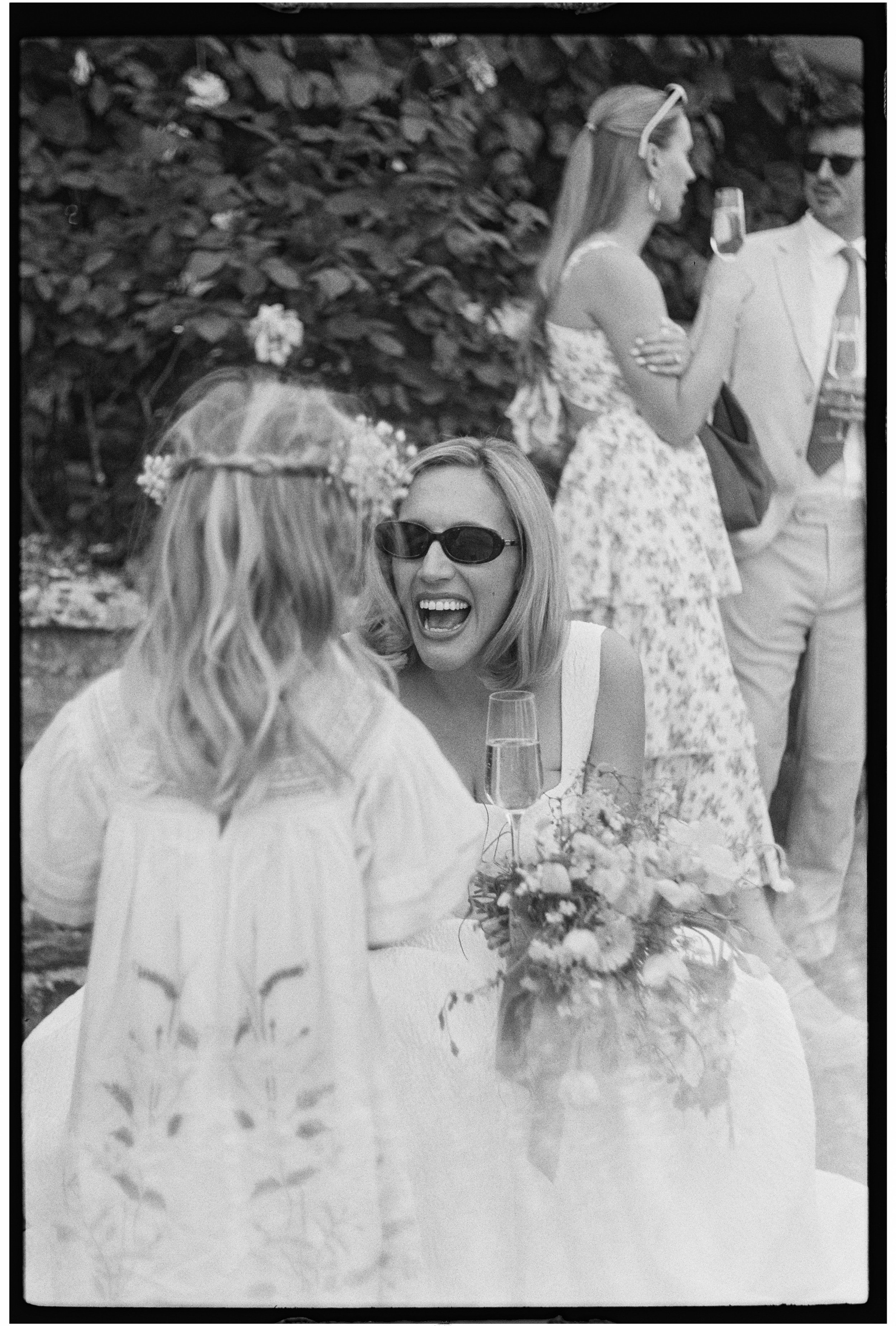 A group of friends at a wedding reception, with a woman in the foreground wearing sunglasses and a white dress, smiling and holding a glass of wine, while another woman and man are in the background, standing with drinks and chatting outdoors.