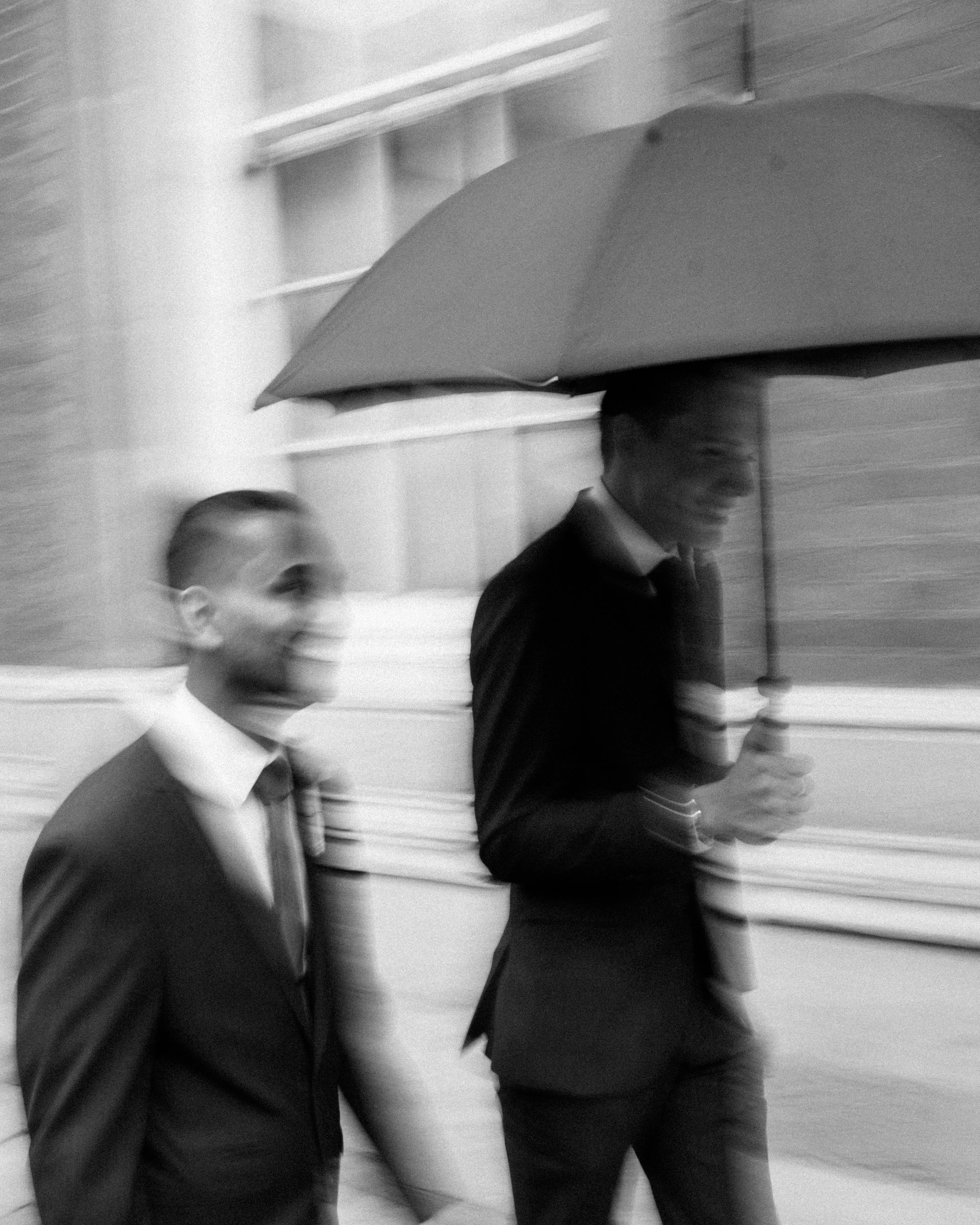 Three people walking together, two men and one woman. The woman is holding an umbrella and wearing glasses, with a smile on her face. The photo is in black and white.