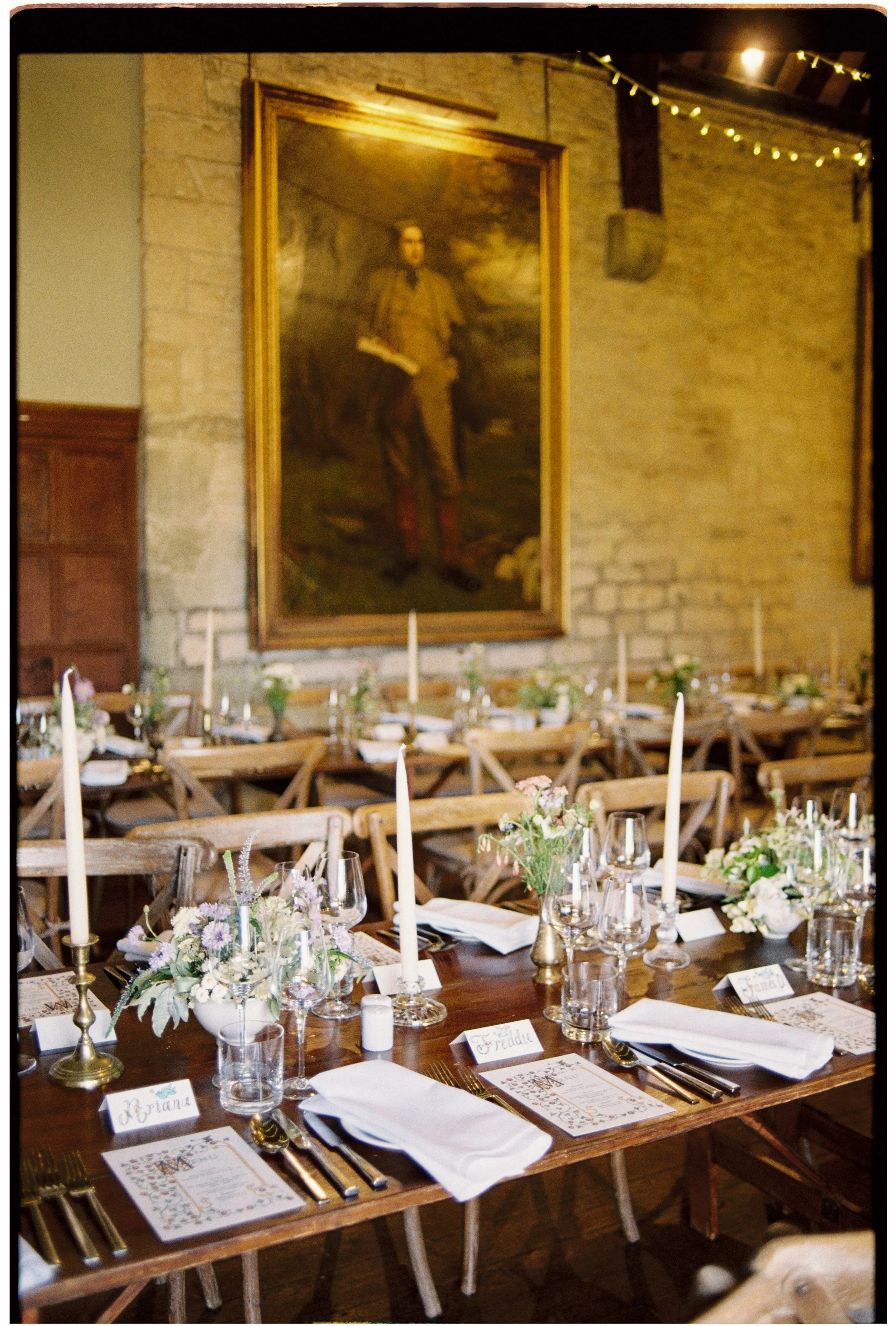 A decorated dining table set for a formal event, with floral centerpieces, candles, wine glasses, and place cards, in a rustic venue with exposed brick walls and a large portrait painting hanging on the wall.