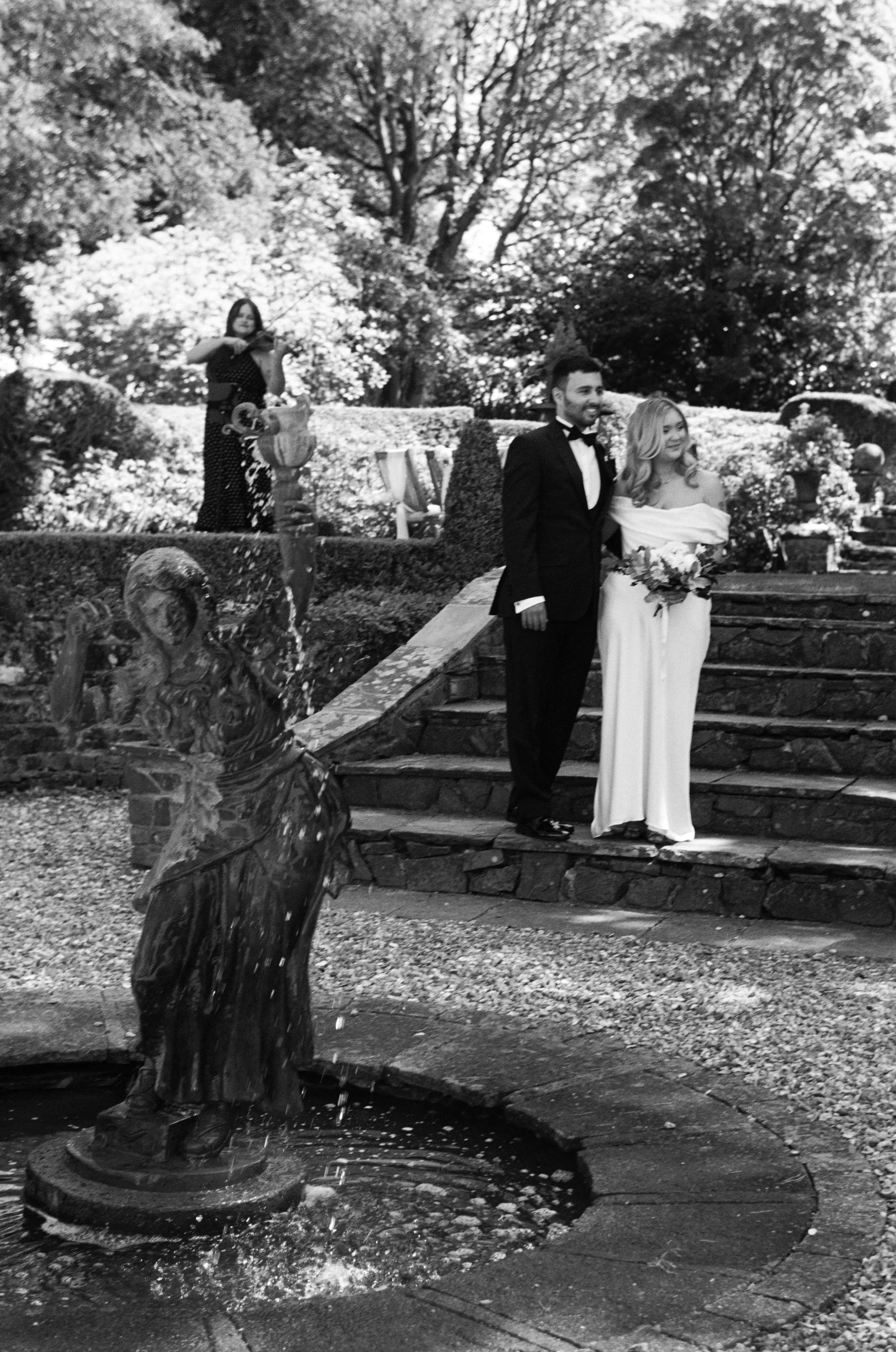 A black-and-white photograph of a fountain with a sculpture of a girl pouring water, surrounded by plants and trees. Two people, a man in a tuxedo and a woman in a wedding dress, stand on the steps behind the fountain.