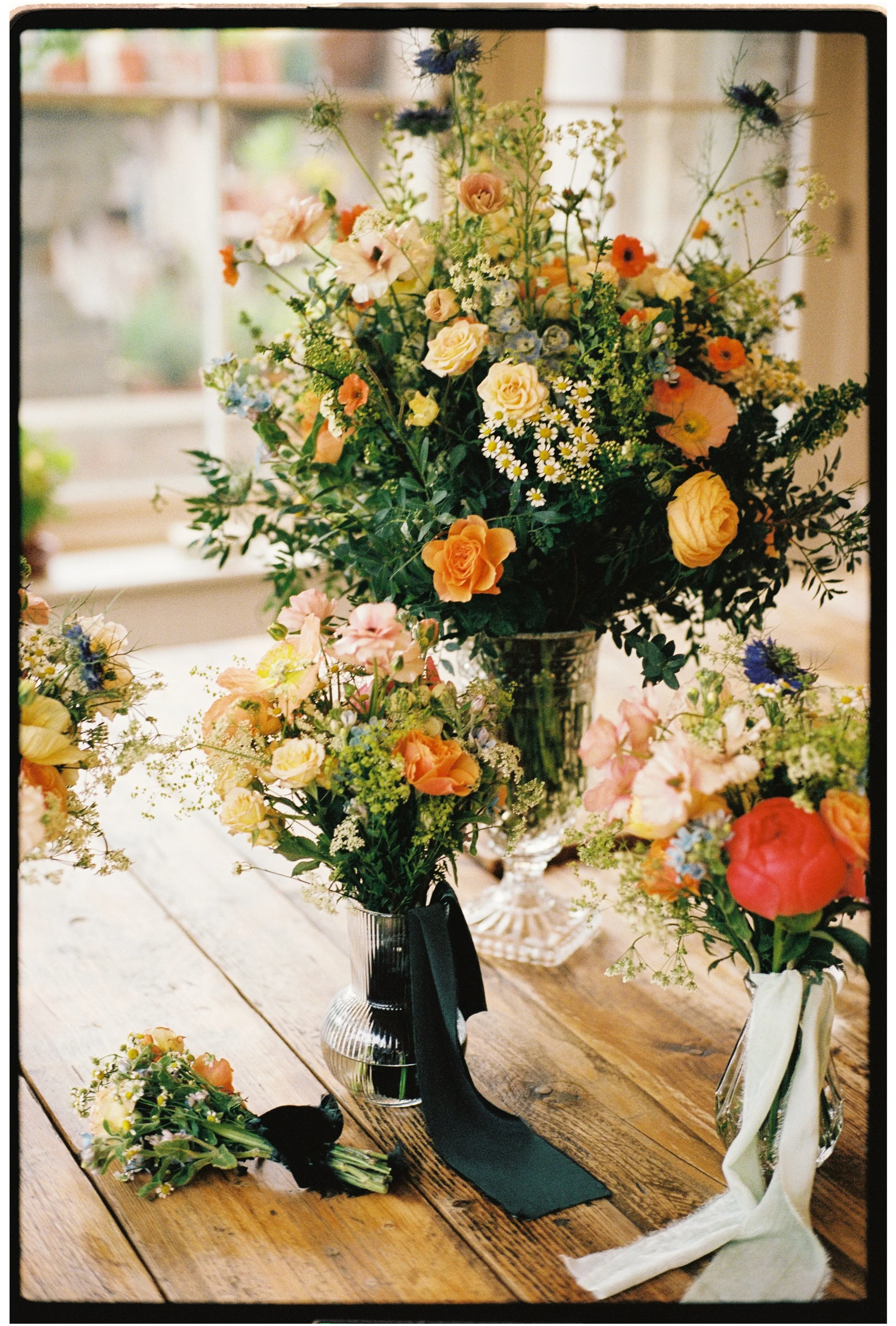 A floral arrangement with yellow, orange, and pink flowers in a glass vase, placed on a wooden table, with additional small bouquets and a white cloth nearby.