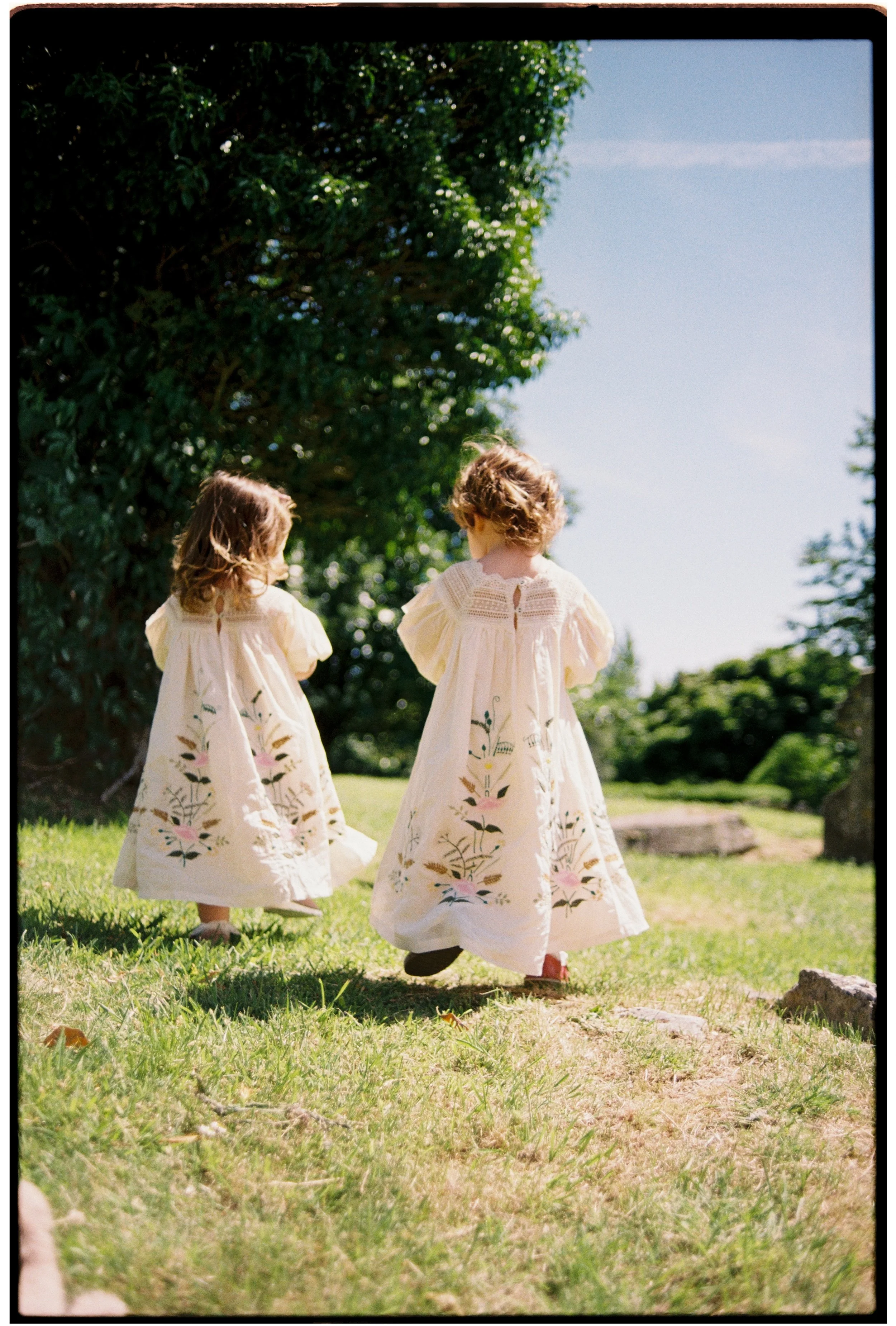 Two young girls in cream-colored dresses with floral embroidery walking outdoors on a sunny day, near a large tree with lush green foliage.