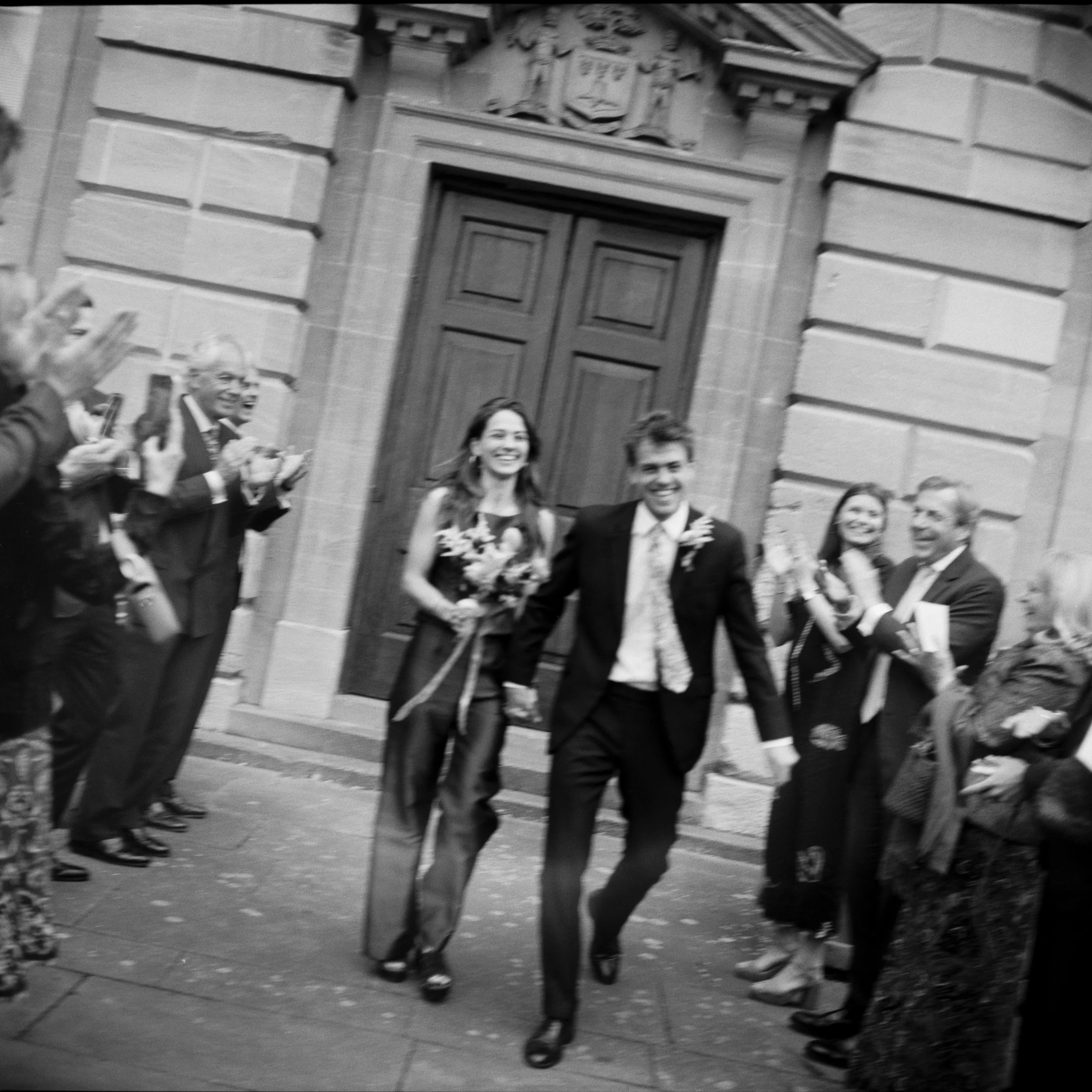 Black and white photo of a group of smiling people in formal attire walking past a historic building with ornate architectural details, including a large wooden door and decorative stonework.