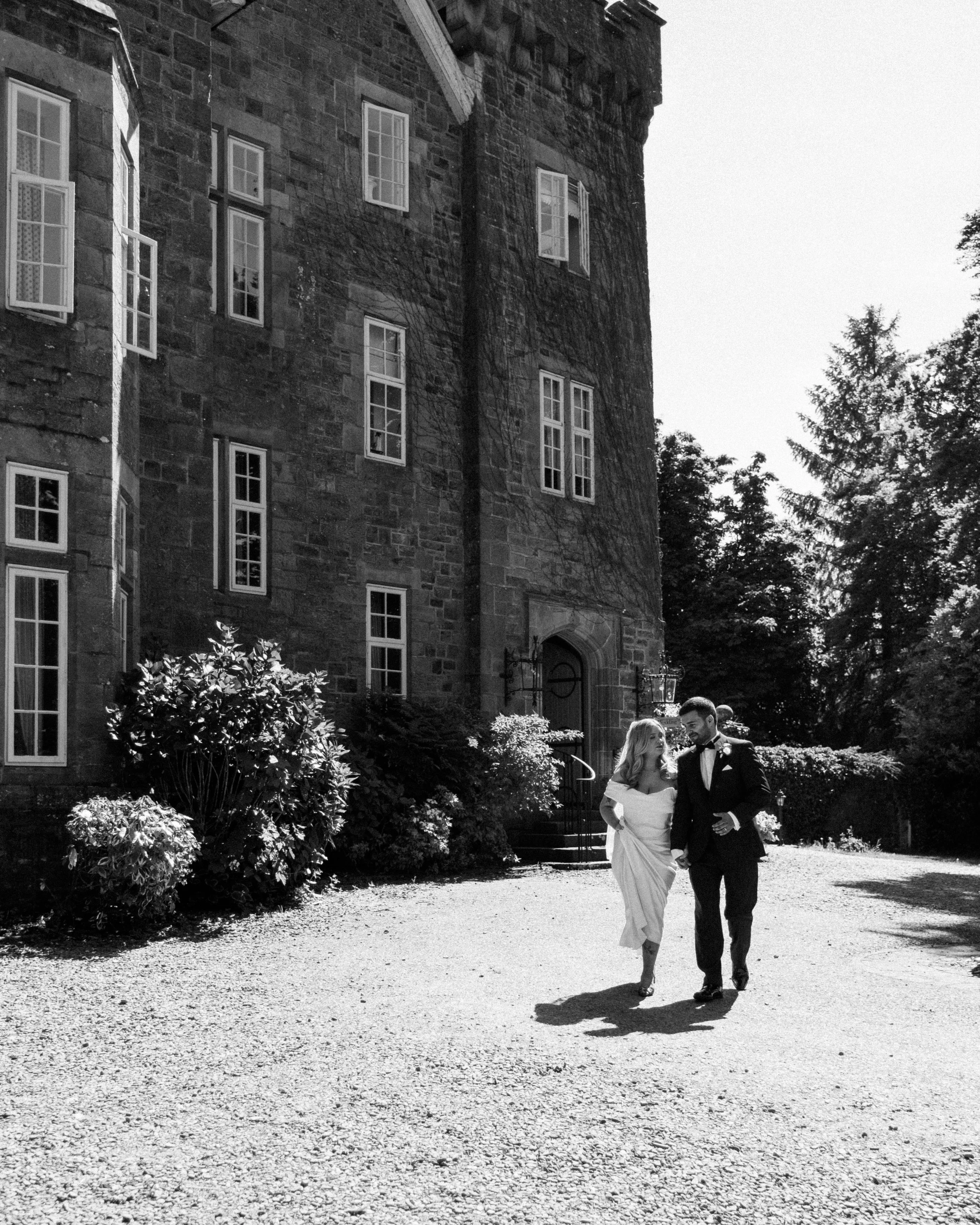 A bride and groom walking together outside a stone building with multiple windows and trees in the background.