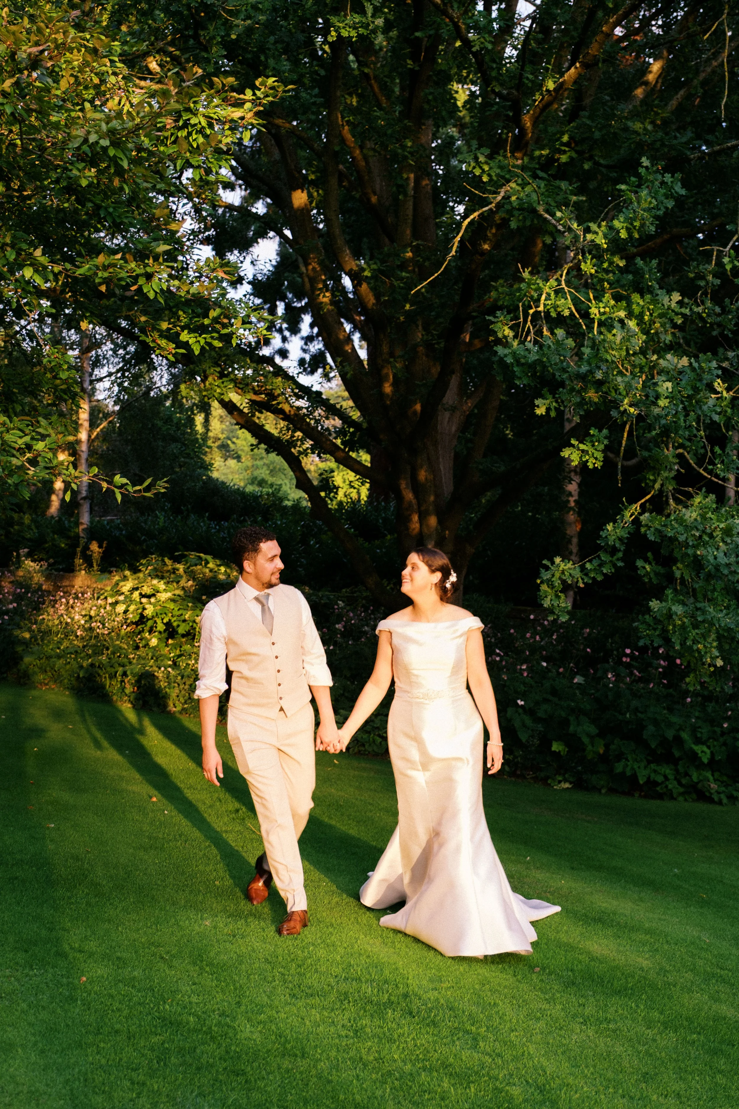 A bride and groom walking hand in hand outdoors in a garden with lush green grass and trees during the evening.