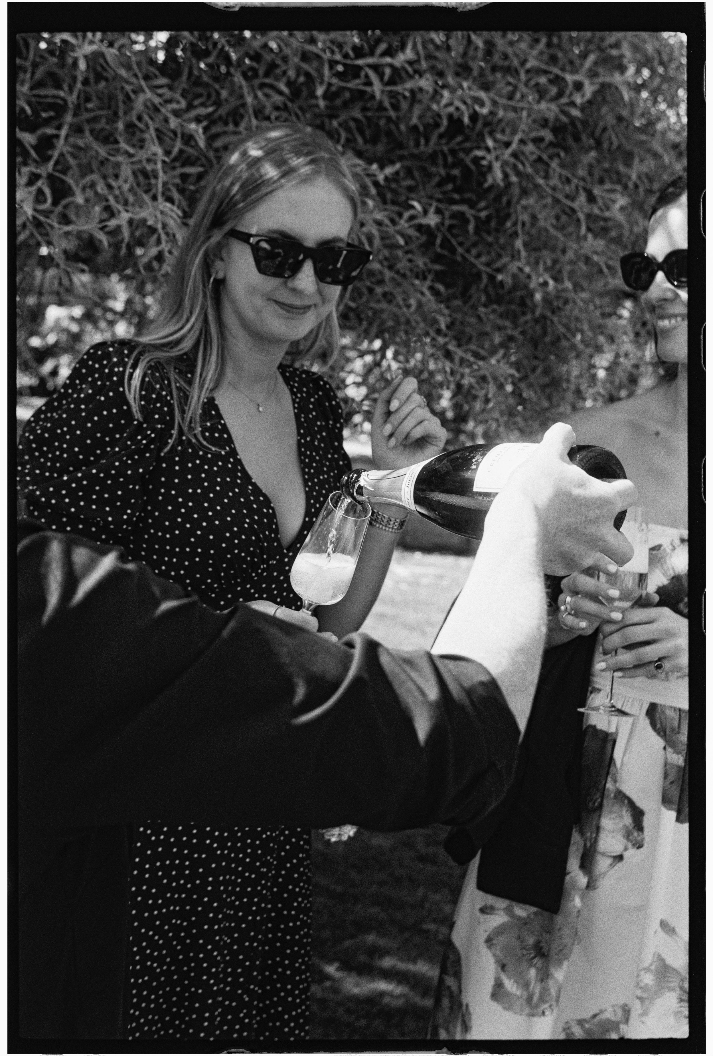 Two women wearing sunglasses at an outdoor gathering, with one pouring champagne into a glass and the other smiling.