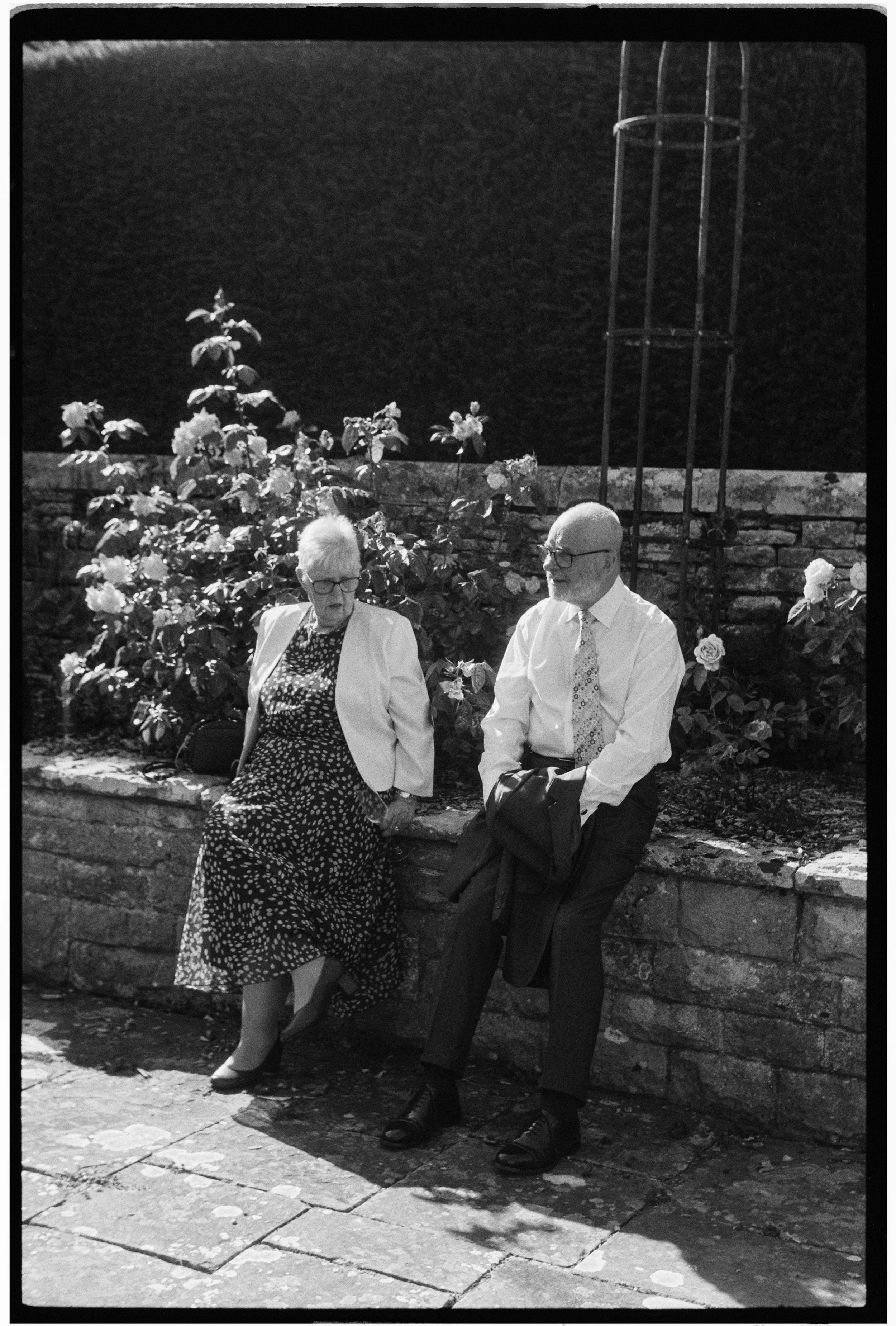 An elderly woman and man sitting on a stone wall in a garden, engaged in conversation. The woman is wearing glasses, a polka dot dress, and a white jacket, while the man is dressed in dark pants, a white shirt, tie, and glasses. There are blooming fl