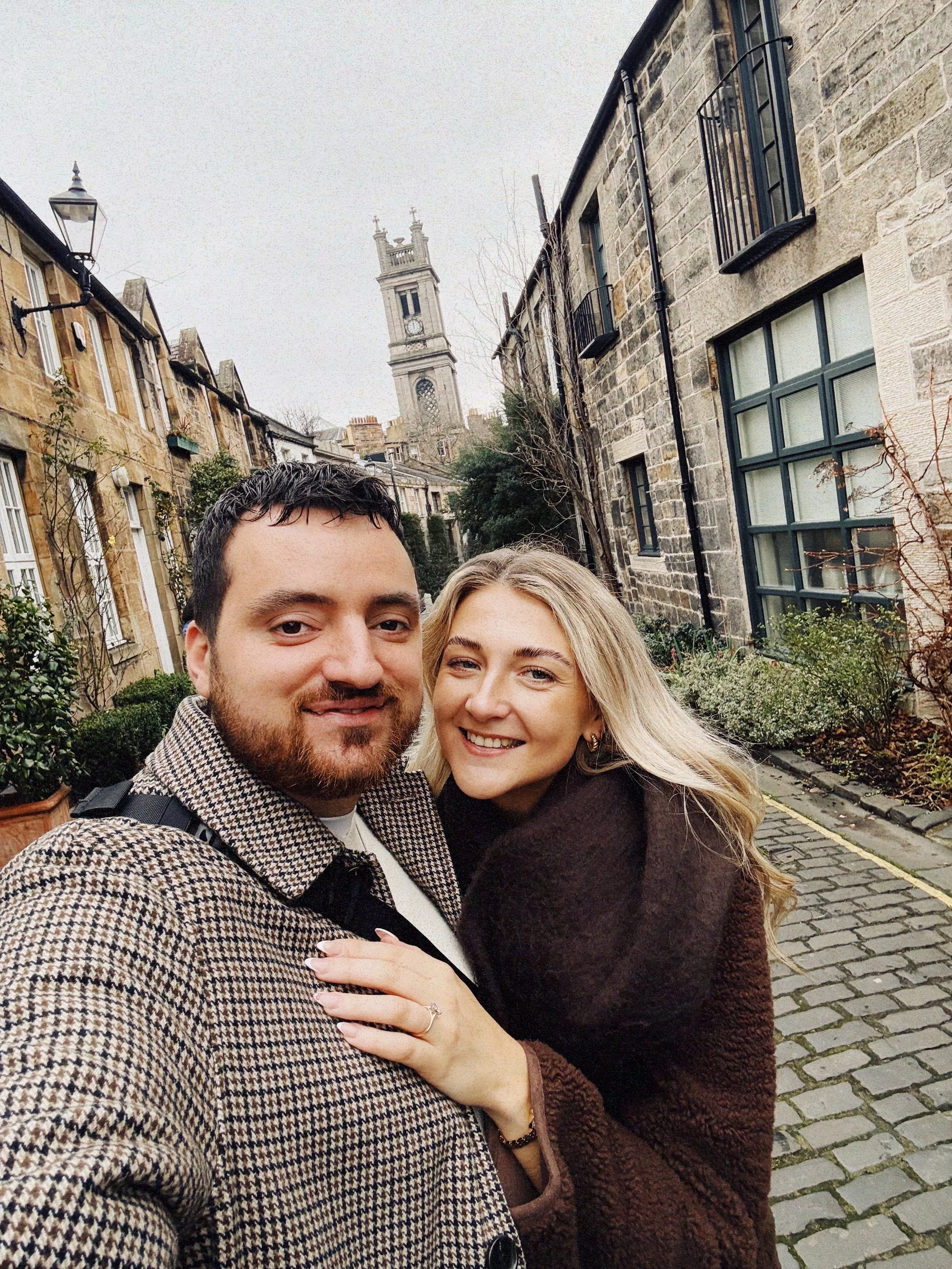 Close-up selfie of a smiling man and woman outdoors on a cobblestone street with historic buildings and a clock tower in the background. The man has dark hair and a beard, wearing a checkered coat, and the woman has long blonde hair, wearing a dark brown coat, with her hand on the man's chest showing an engagement ring.