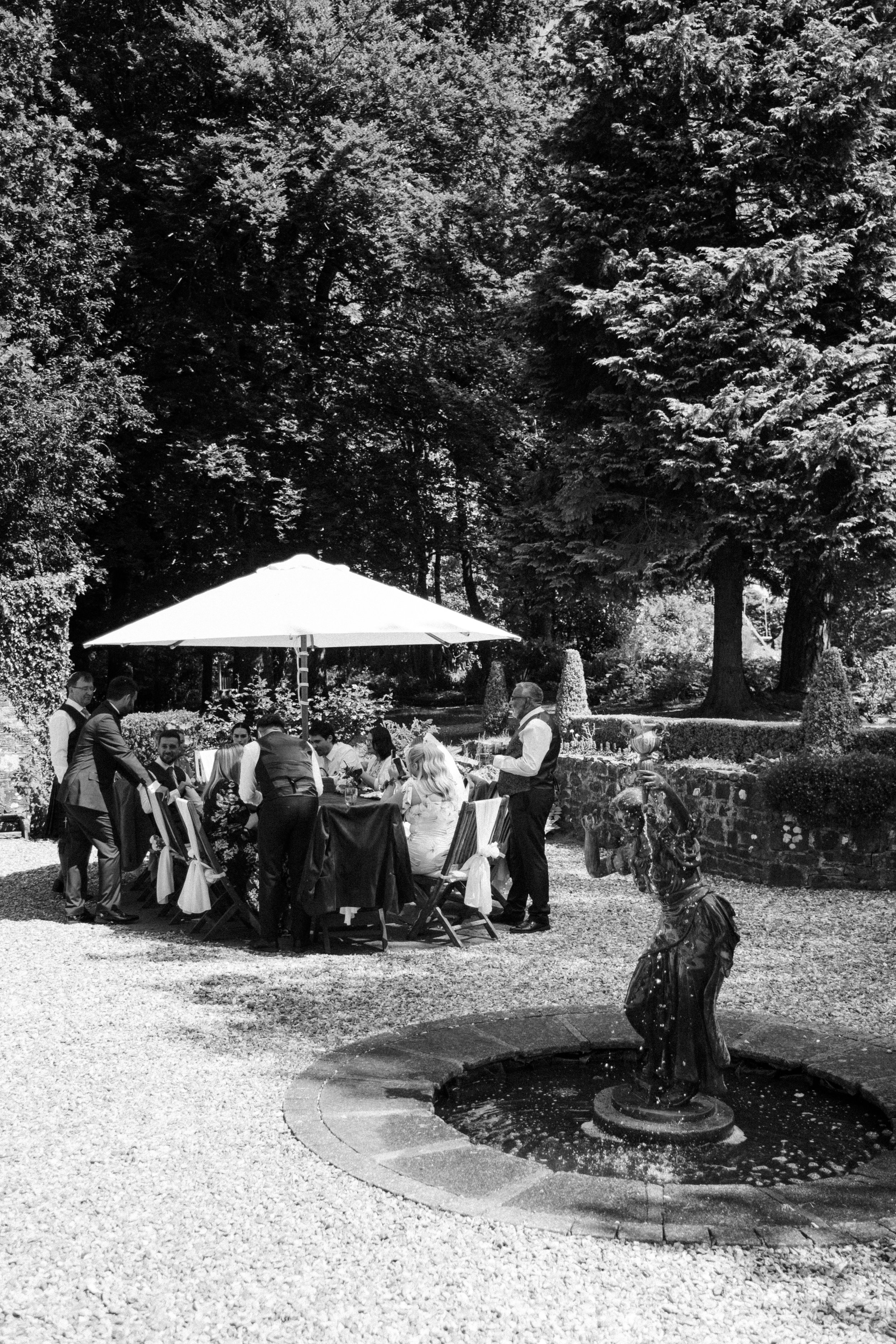 Outdoor gathering with people seated at a table under a large umbrella, near a decorative fountain with a horse statue in a garden surrounded by trees.