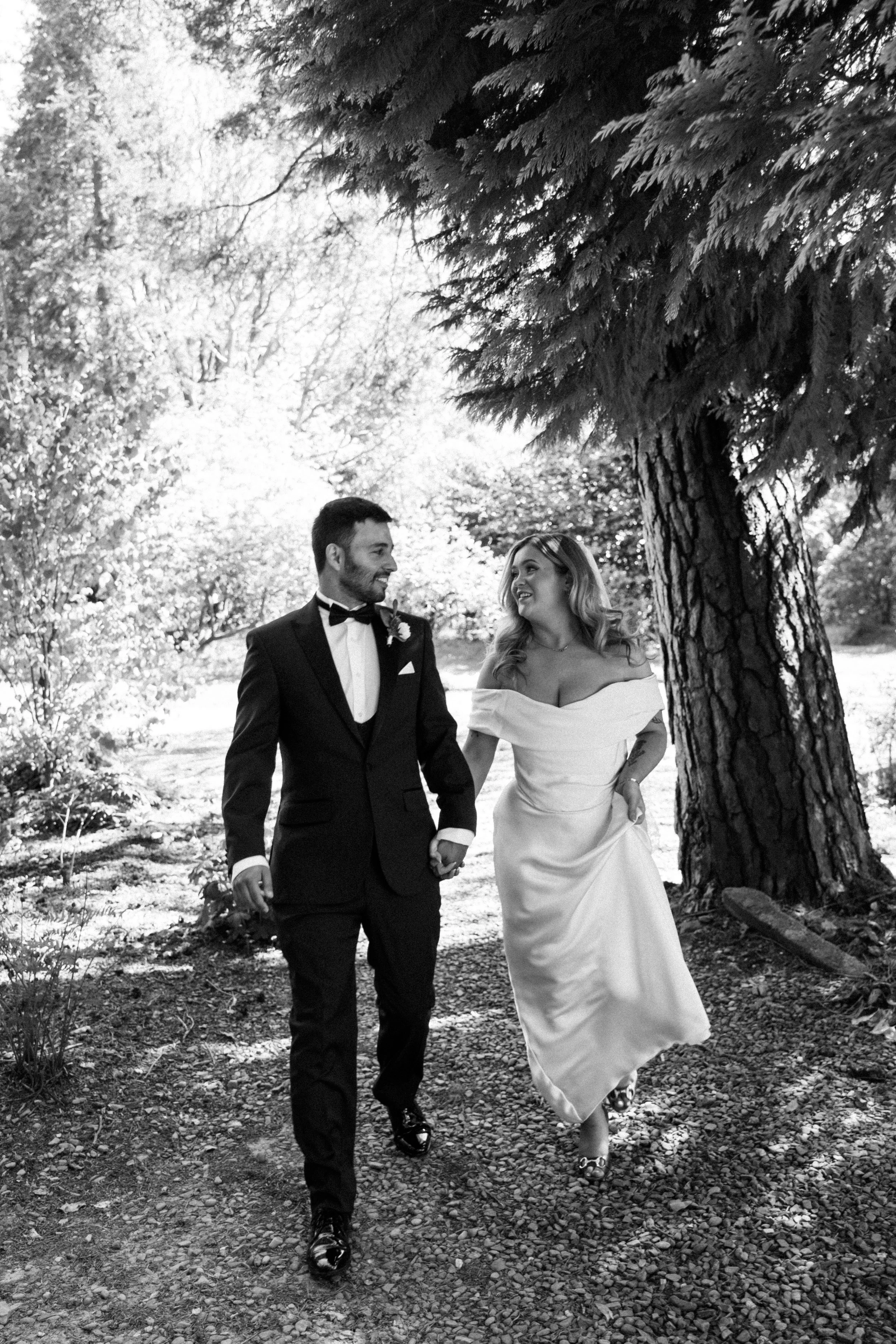 A black and white photo of a bride and groom walking hand in hand outdoors under a large tree, smiling at each other.