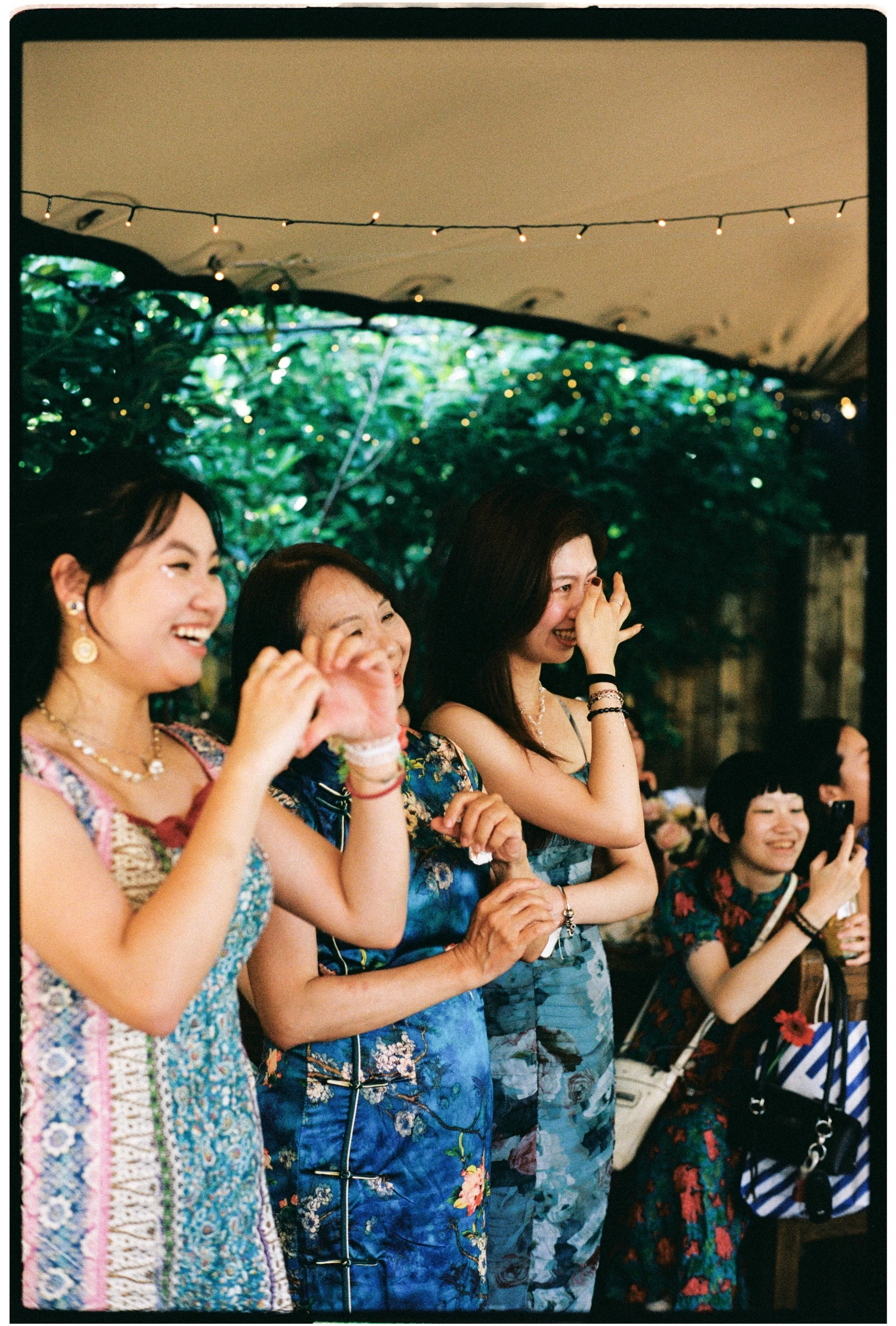 A group of women wearing colorful dresses are gathered, smiling and laughing, at an outdoor event decorated with string lights and lush greenery.