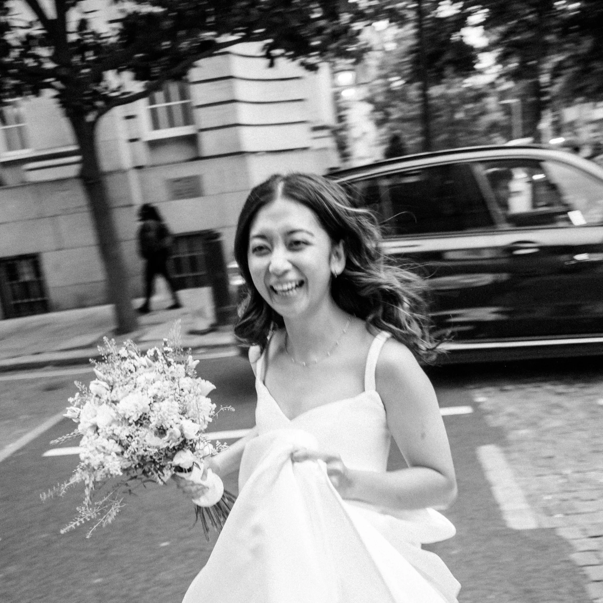A woman with long hair holding a bouquet of flowers, smiling, with cars and trees in the background.