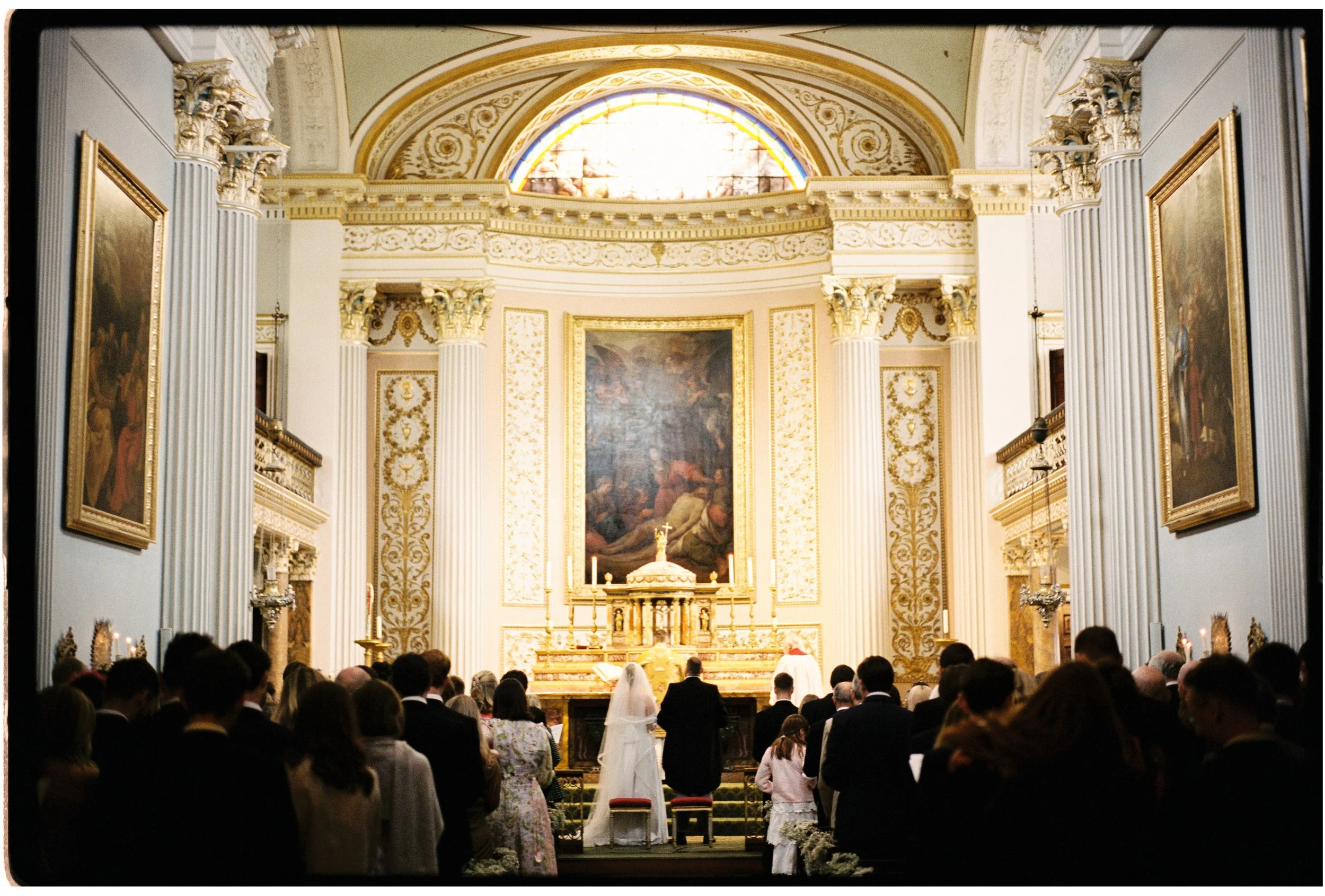 A wedding ceremony taking place inside a grand church with ornate gold and white decorations, tall columns, and large paintings on the walls. The bride and groom stand at the altar facing the priest while guests are seated and watching.