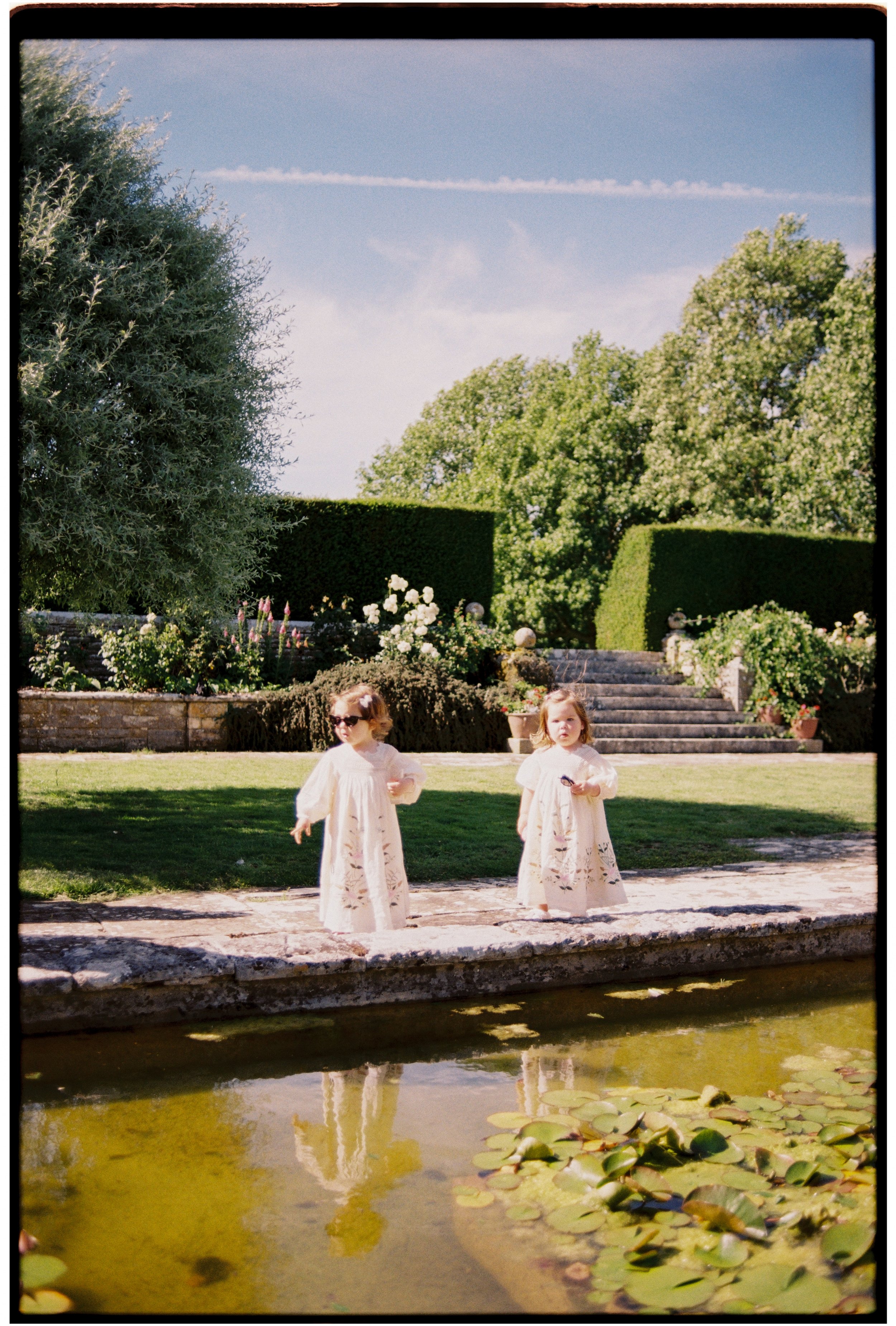 Two young girls in cream-colored dresses with floral embroidery standing by a pond with lily pads, reflecting trees and a blue sky with some trees and bushes in the background.