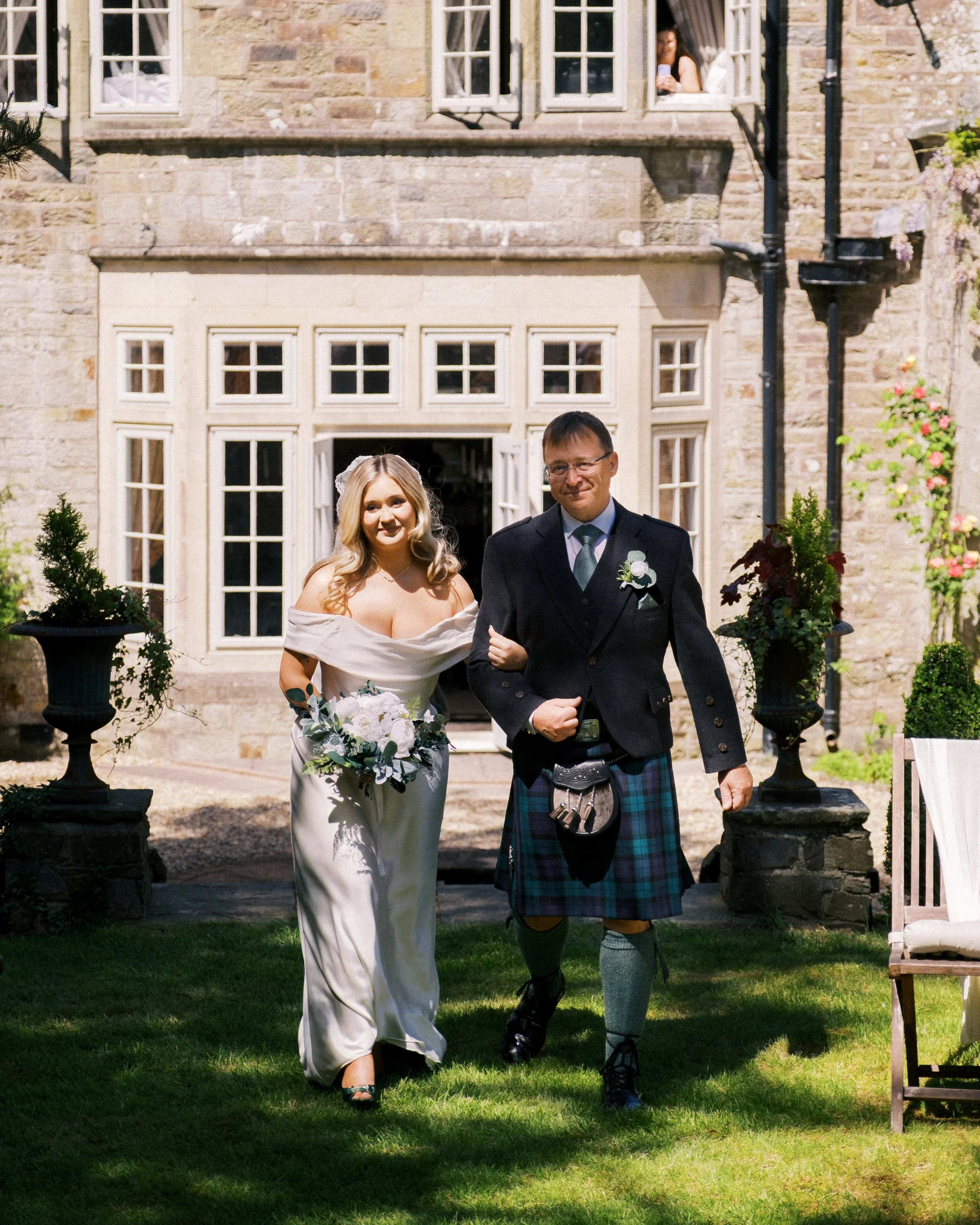 A bride and groom walking outside a stone building, with a woman looking out a window in the background.