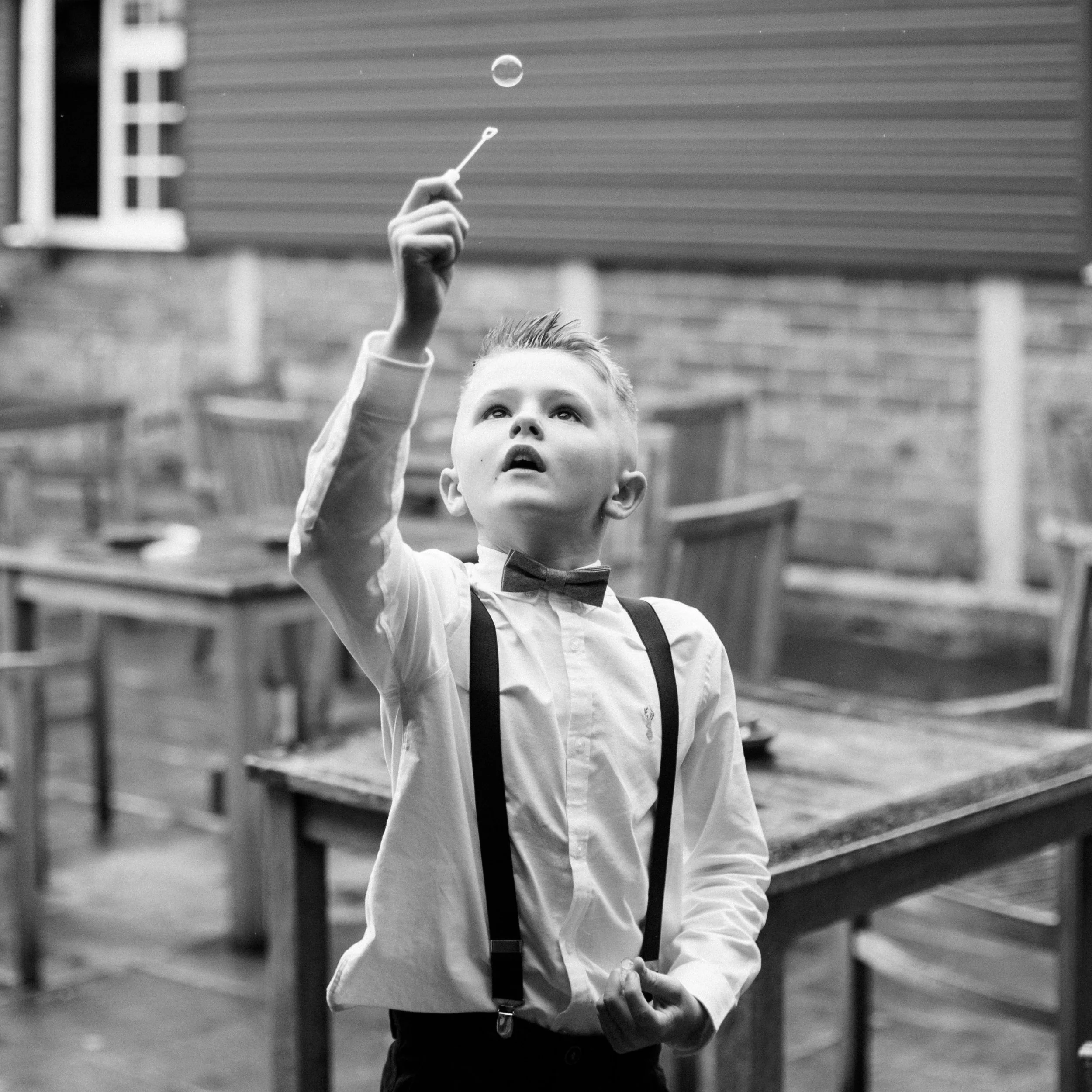 A young boy with short hair, wearing a white shirt and suspenders, is reaching up with his right hand while holding a small doll in his left hand. He is looking up at the moon in the sky. The scene appears to be outdoors in front of a house with horizontal siding.