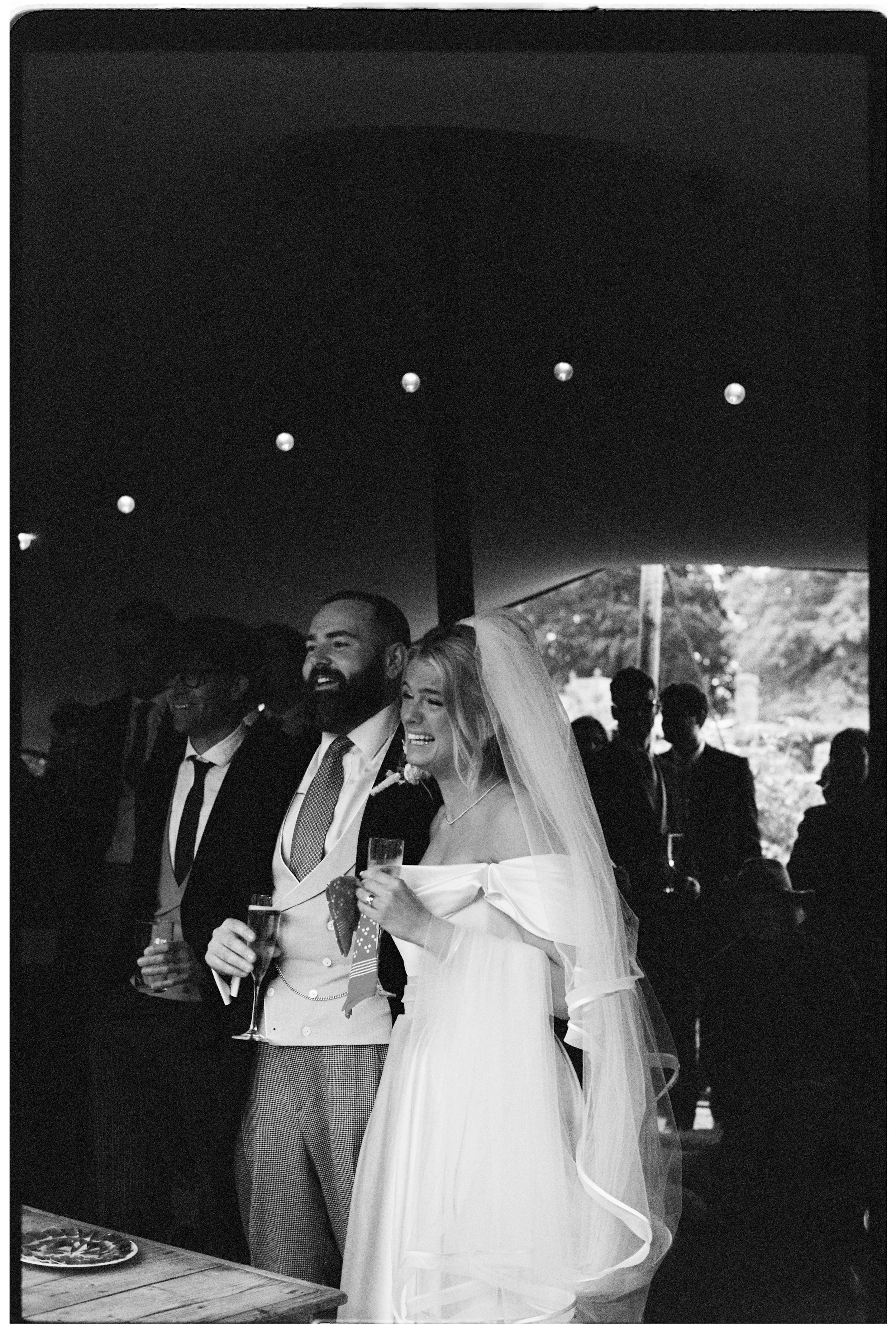 A bride and groom at their wedding reception, smiling and holding drinks, surrounded by guests.