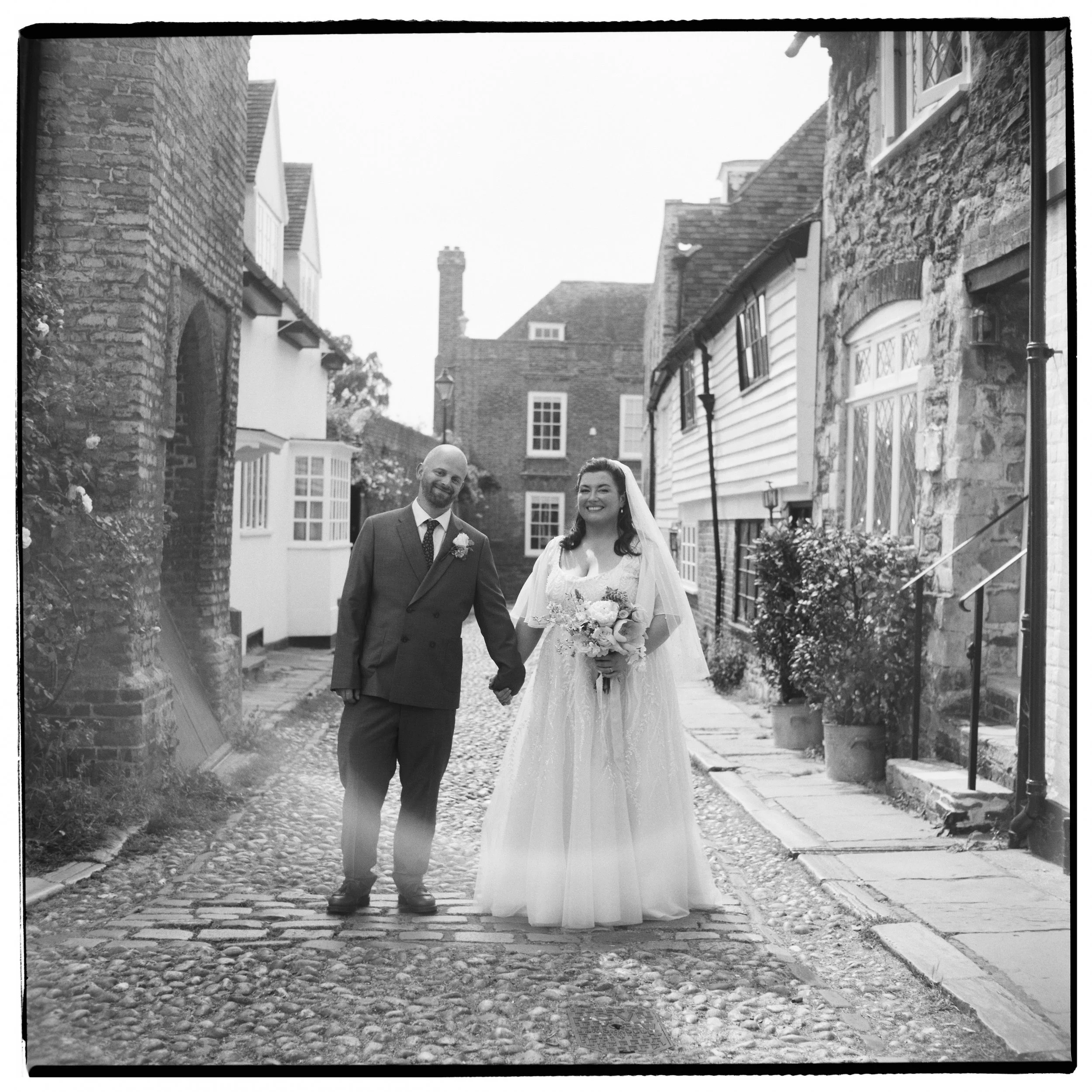 A black and white photo of a bride and groom walking hand in hand down a cobblestone street in a historic neighborhood, with brick and stone houses on either side and chimneys visible in the background.