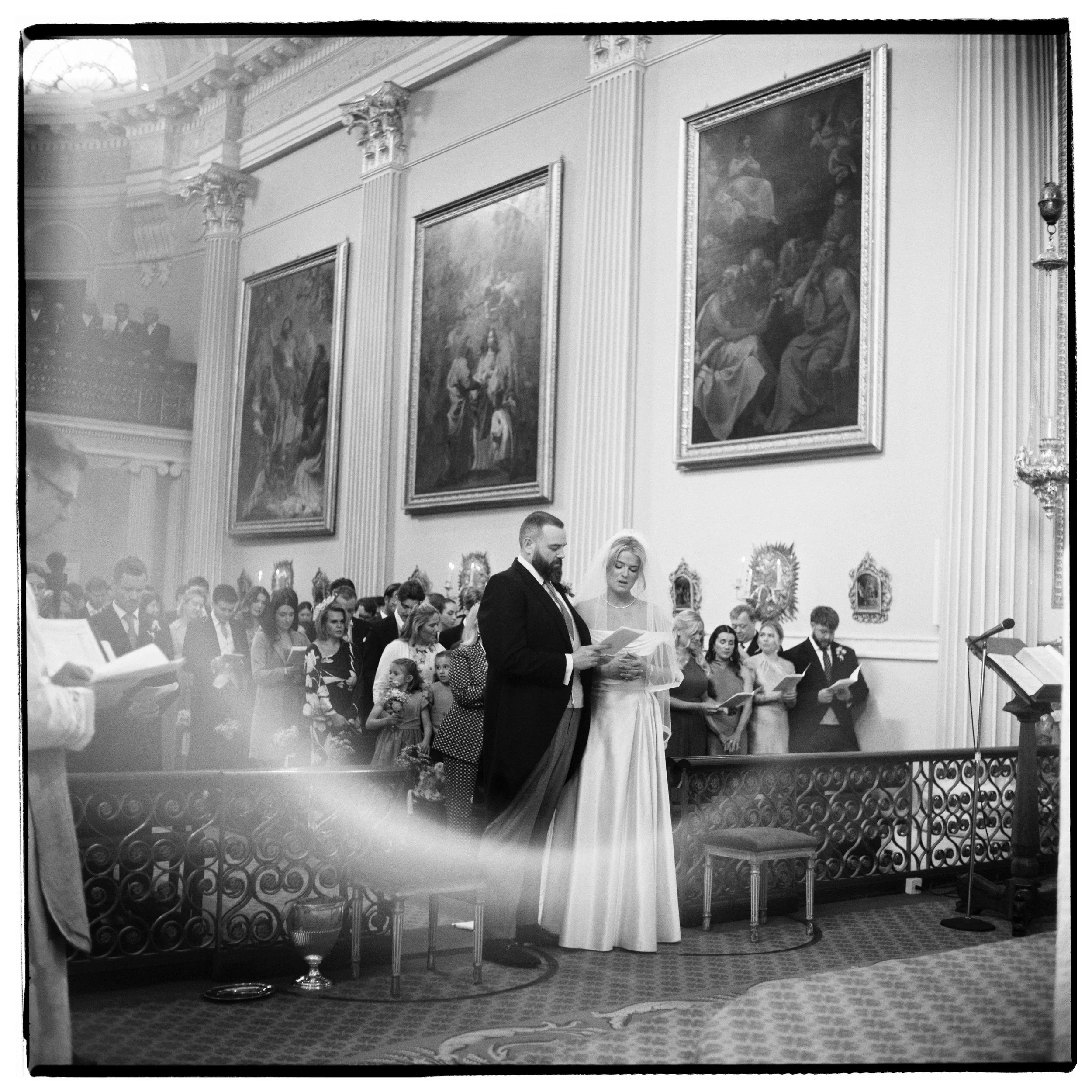 People attending a formal event or ceremony in a grand, ornately decorated hall with large framed paintings on the wall, a decorative staircase, and floral arrangements.