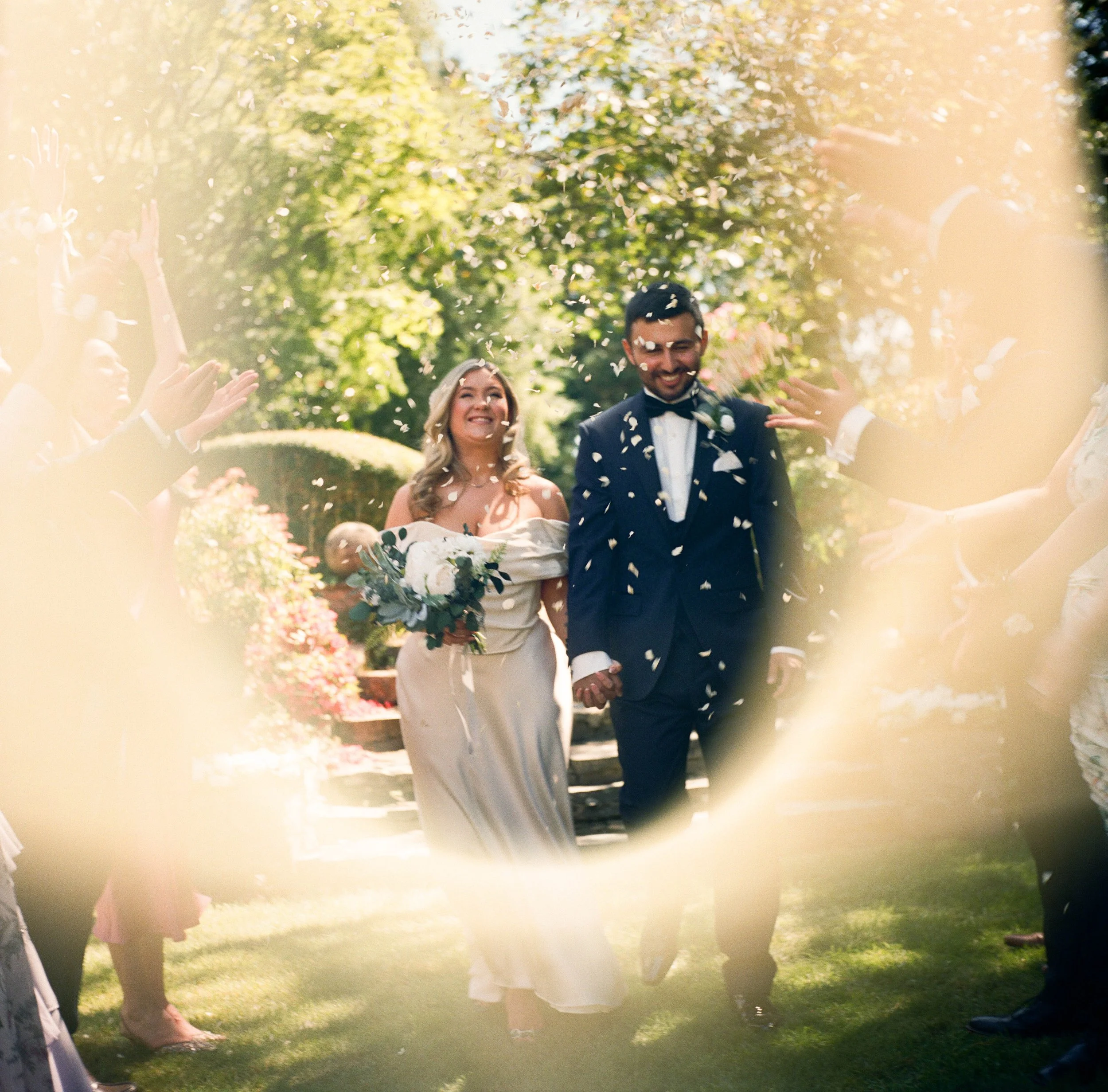 Wedding ceremony outdoors with a bride and groom walking together, surrounded by several guests clapping and throwing flower petals.