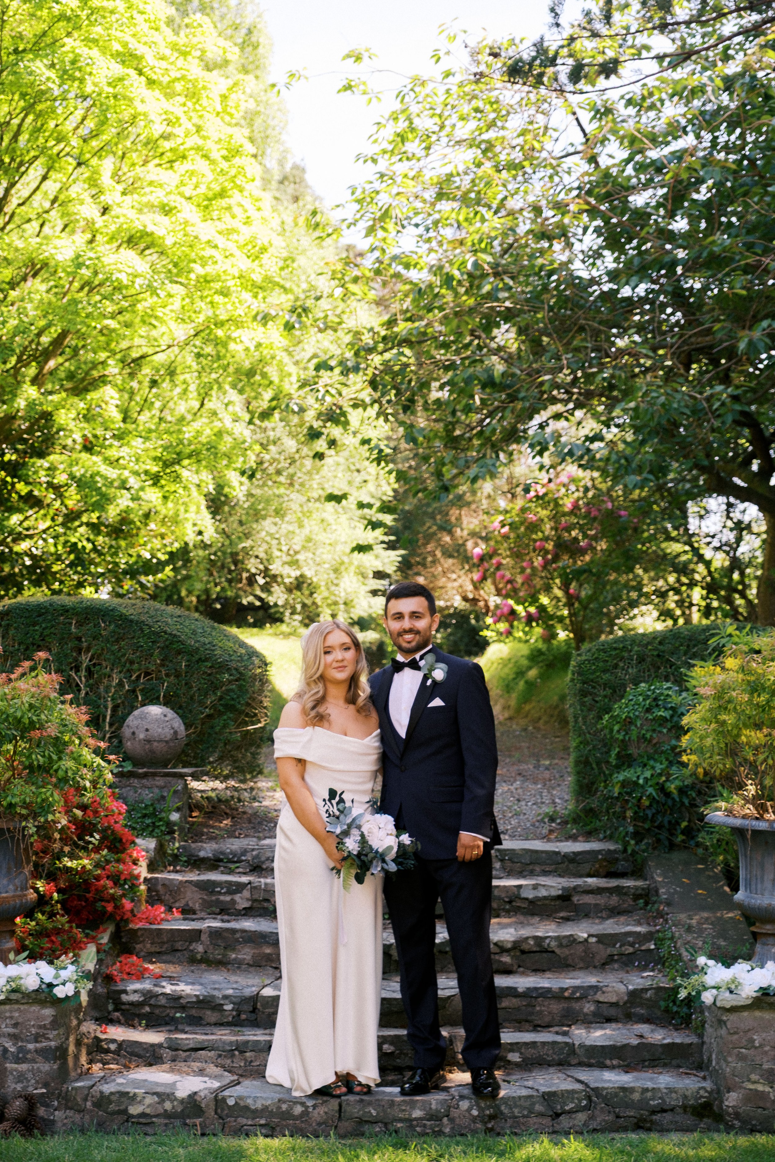 A bride and groom standing on stone steps in a lush garden with green trees and flowering bushes, during a wedding photoshoot. The bride is wearing a strapless, off-the-shoulder white dress and holding a bouquet, and the groom is in a dark tuxedo wit