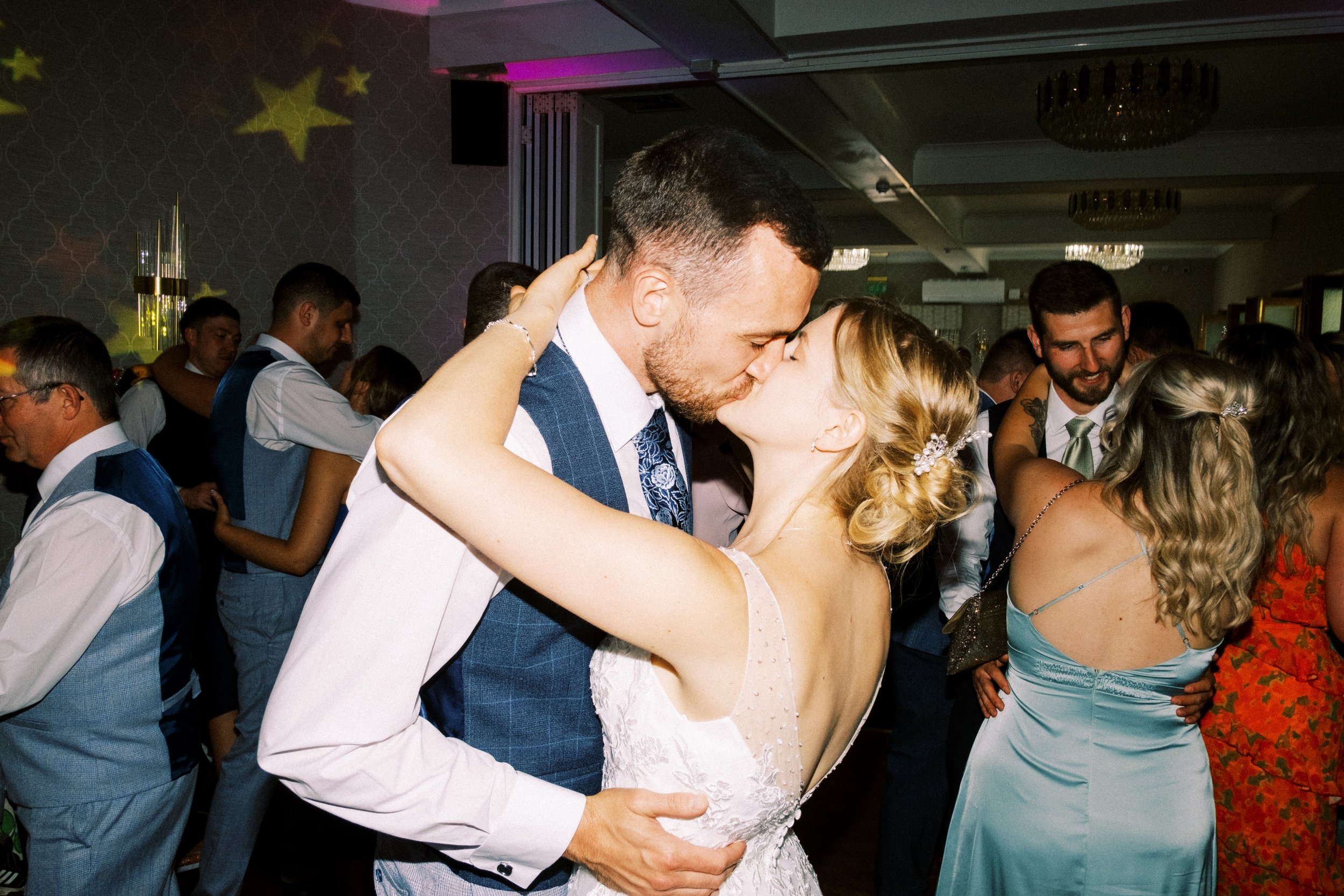 A couple dancing closely, about to kiss, at a wedding reception. The man is wearing a blue suit and the woman a white wedding dress. Other guests are dancing around them in a decorated ballroom.