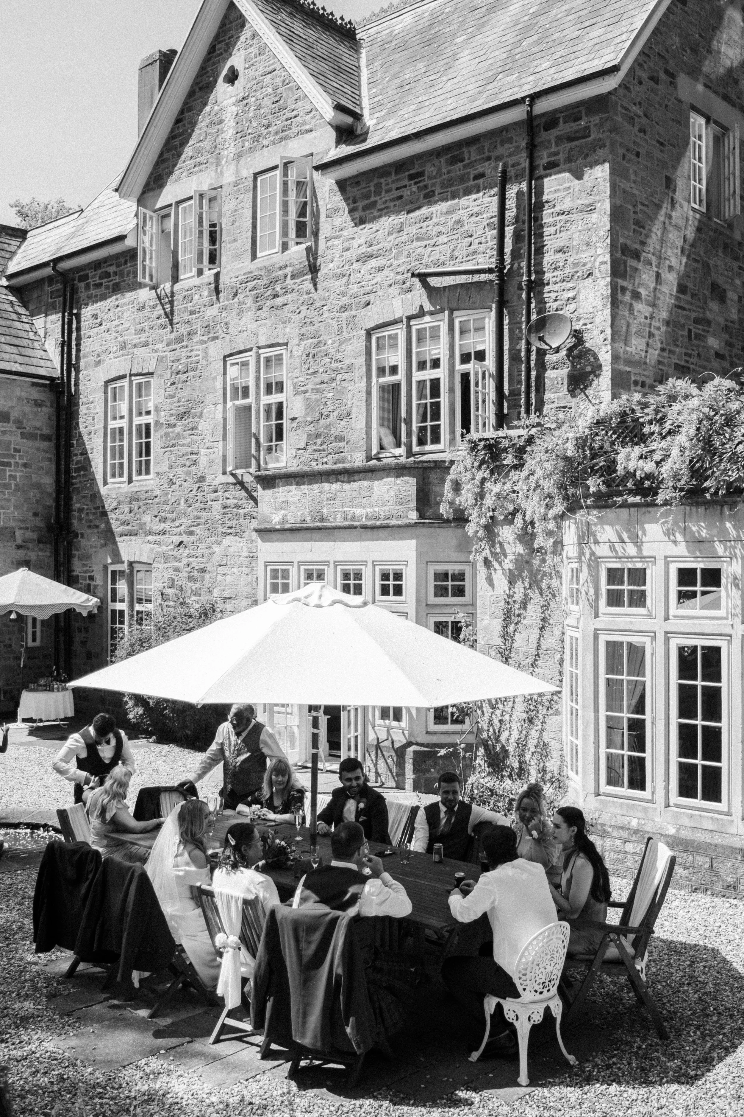 People at an outdoor wedding reception in front of a stone building with multiple open windows. Guests are seated around a long table under umbrellas, enjoying food and drinks.