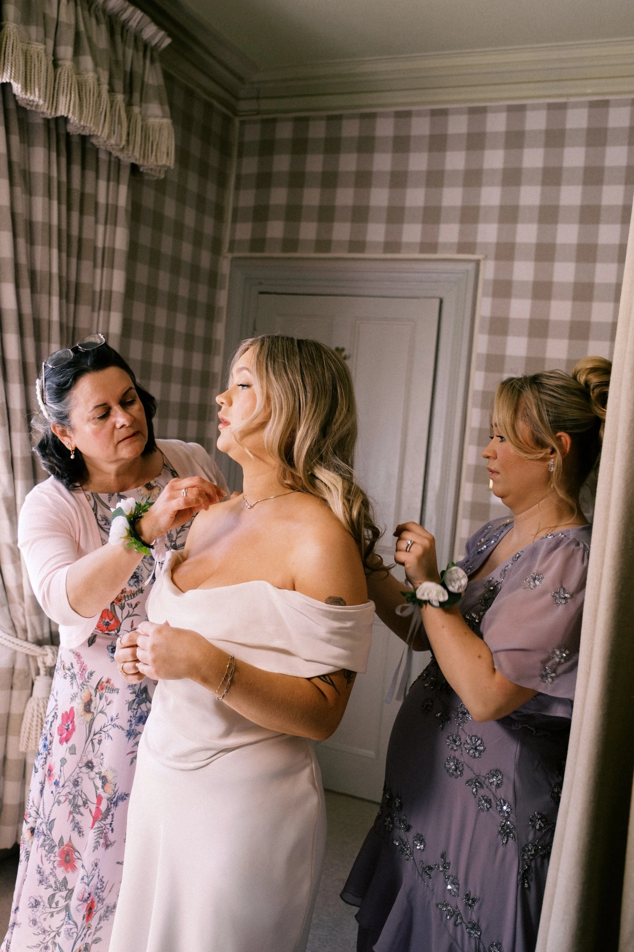 A bride getting dressed with the help of two women in a softly lit room with checkered wallpaper and floral curtains.