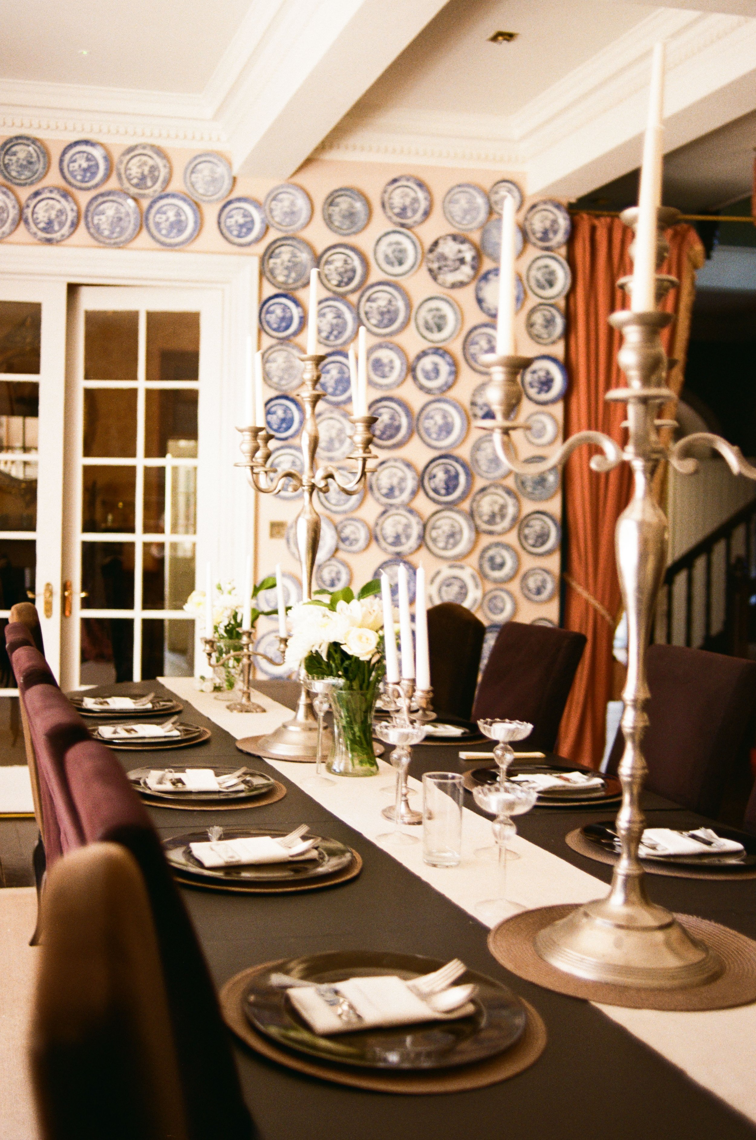A dining room decorated for a formal gathering or celebration, featuring a long table with place settings, silver candelabras, and a centerpiece of white flowers in a glass vase. The background displays a wall adorned with decorative blue and white p