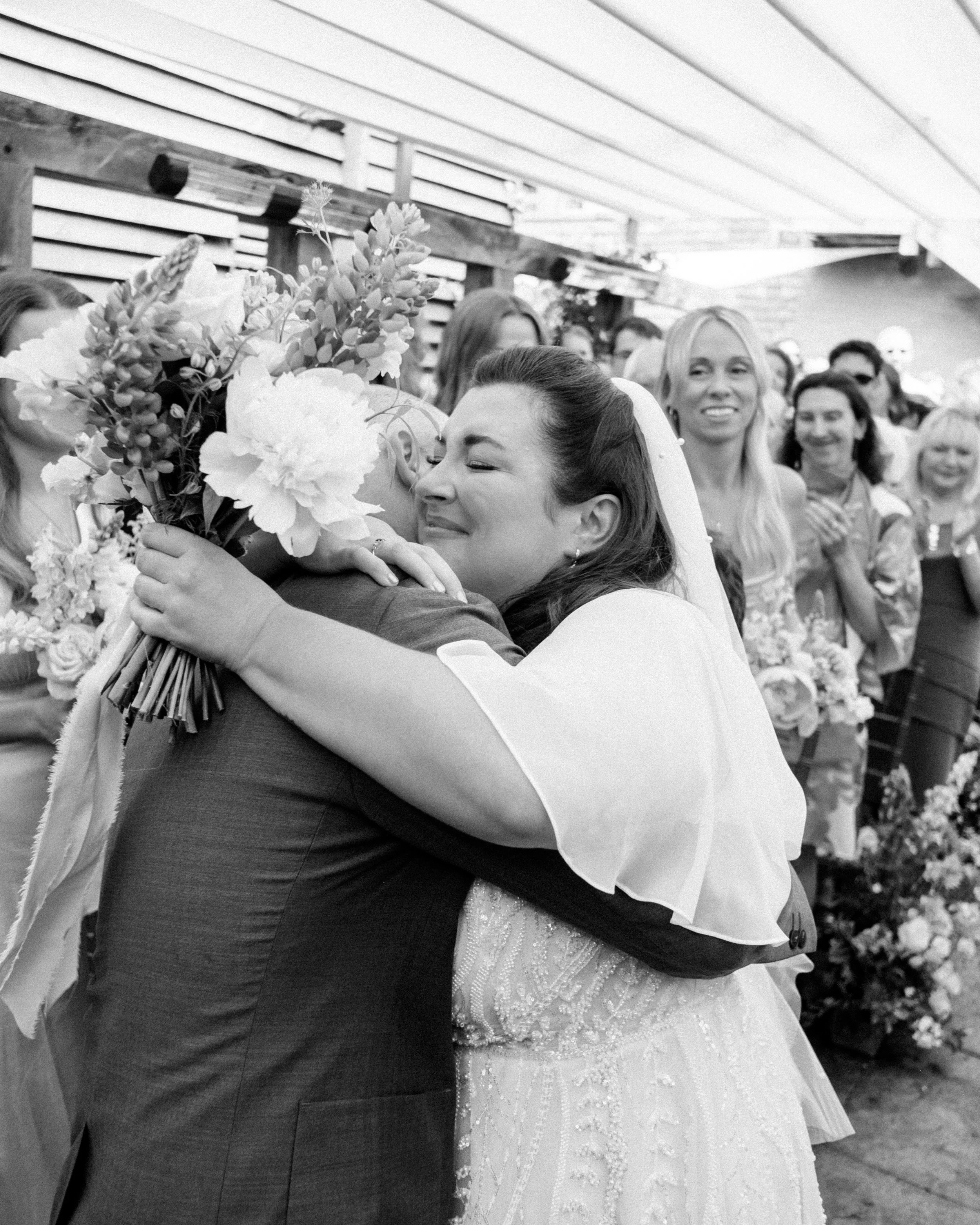 A bride and groom embrace, with the bride holding a bouquet of flowers. In the background, women are standing and smiling. The scene appears to be at a wedding celebration.