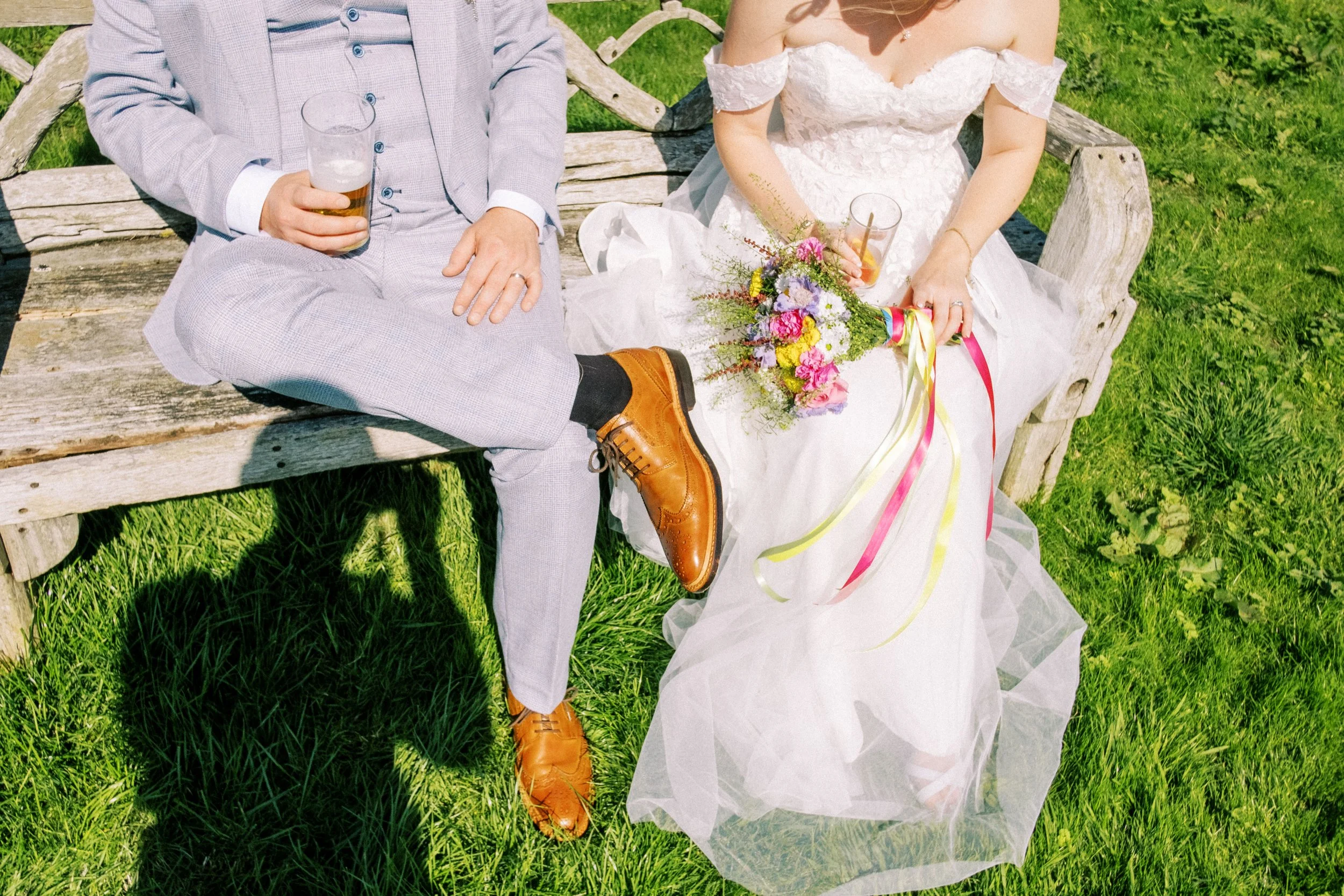 A bride and groom sitting on a wooden bench outdoors, with the bride holding a bouquet of flowers and the groom holding a glass of beer.