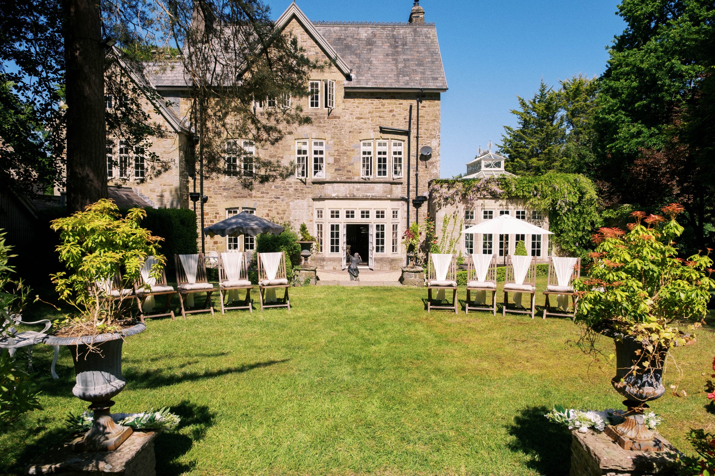 A stone mansion with many windows and a sloped roof, surrounded by green trees and a well-kept lawn. In the foreground, there are chairs and tables with umbrellas, set up in a garden area.