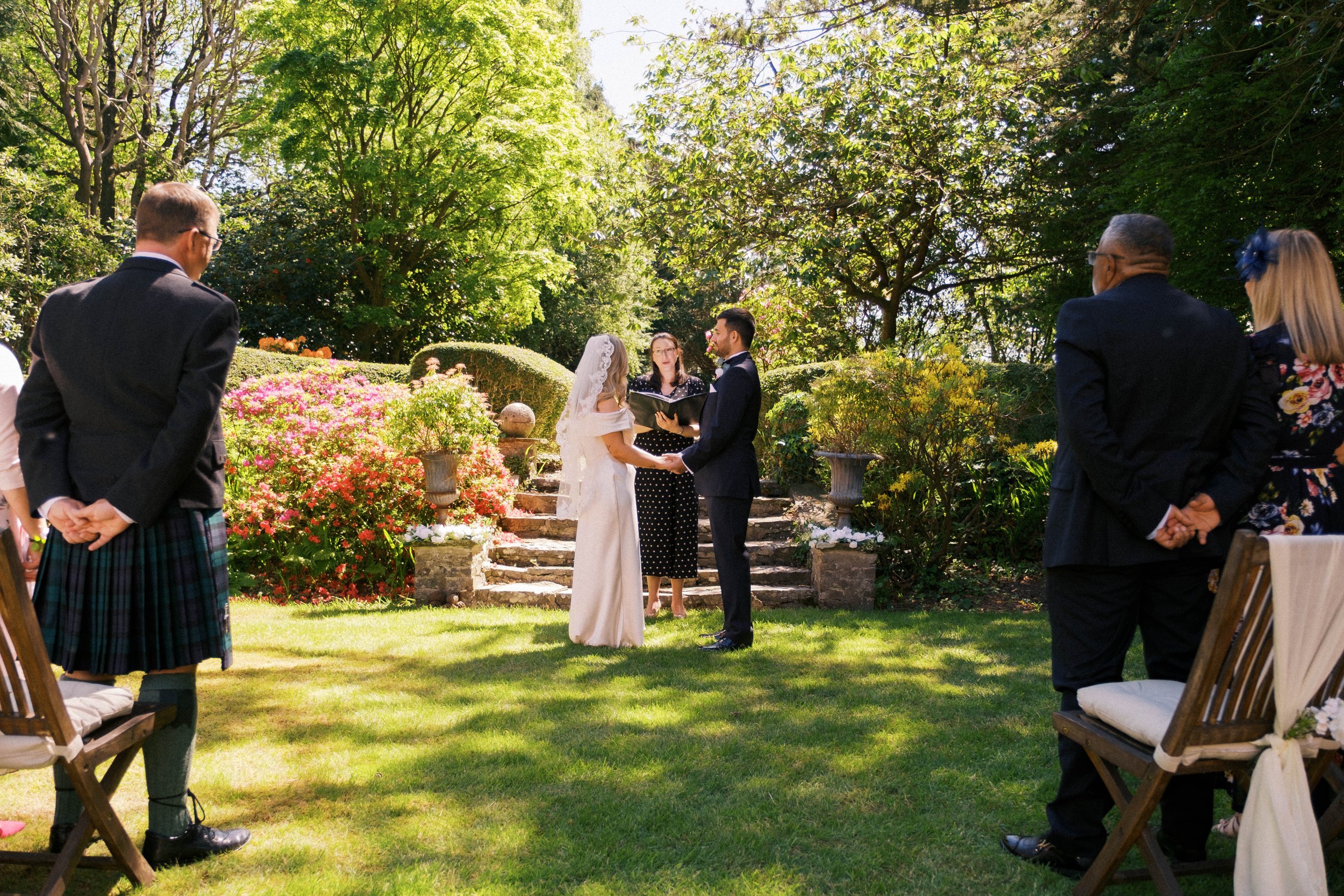 A wedding ceremony outdoors in a lush garden with the bride and groom holding hands, surrounded by seated guests and vibrant greenery.