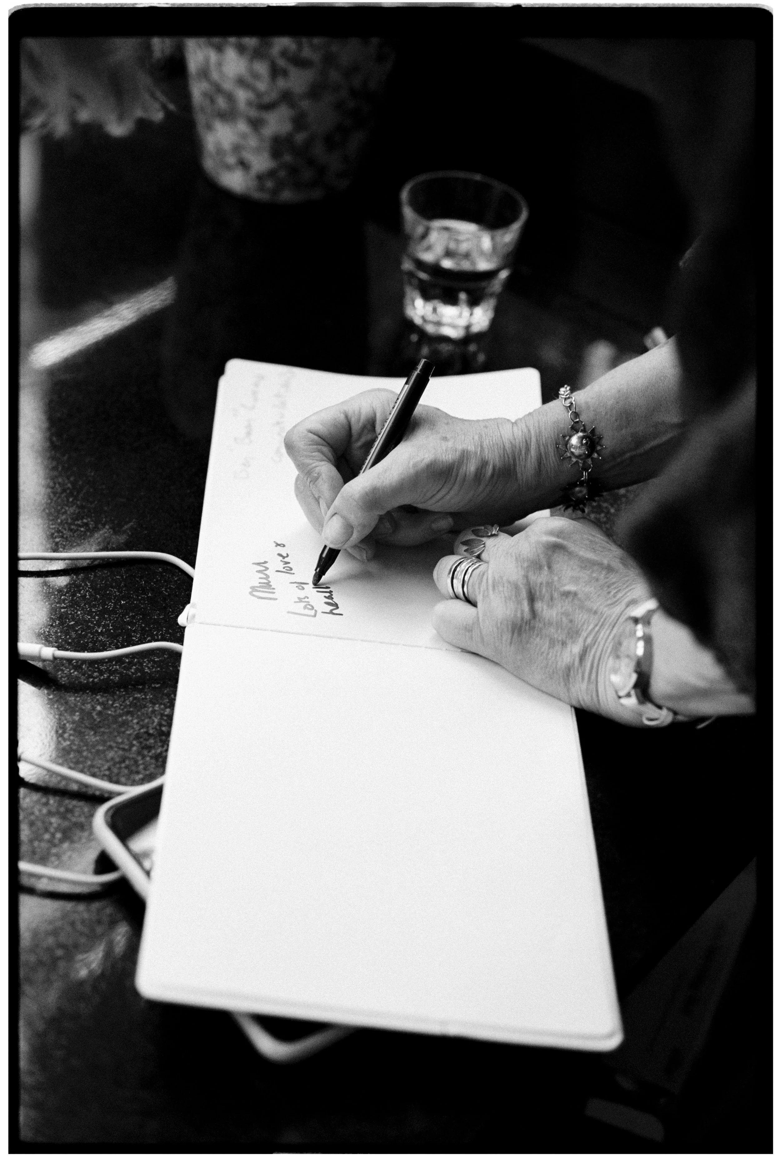 A person's hand holding a pen and writing in a notebook, with a glass of water and a flower pot in the background, on a dark table.