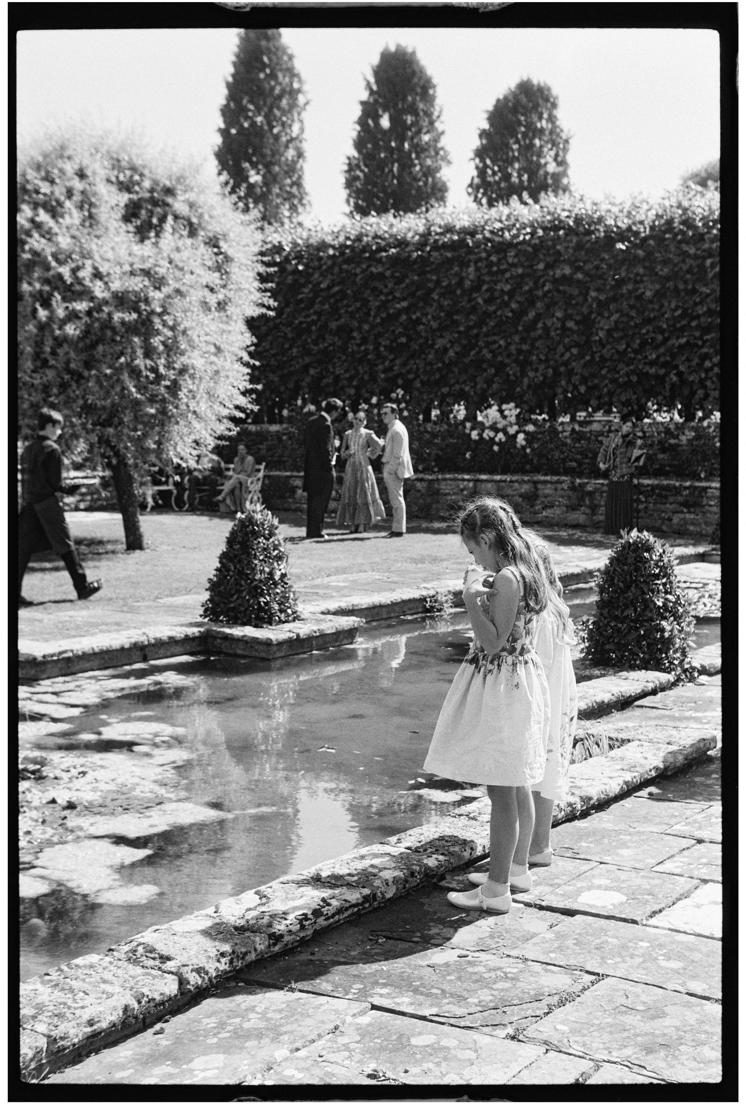 A vintage black-and-white photograph of a young girl in a dress standing by a small pond, with other people in the background and lush trees in a park setting.