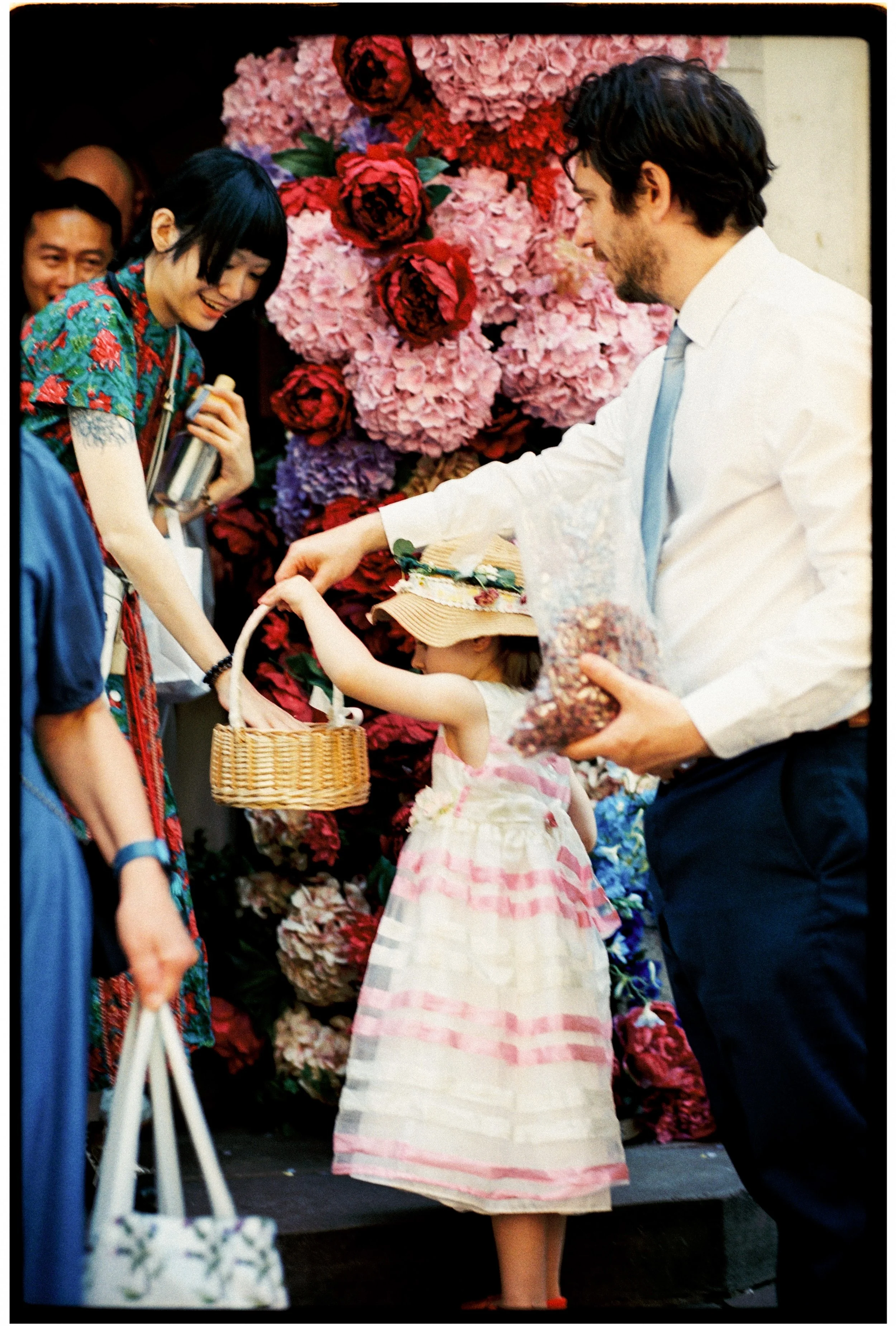 A woman in a colorful dress handing a basket to a young girl in a striped dress, with a man in a white shirt and tie smiling nearby at a flower stand with large pink hydrangeas and red roses.