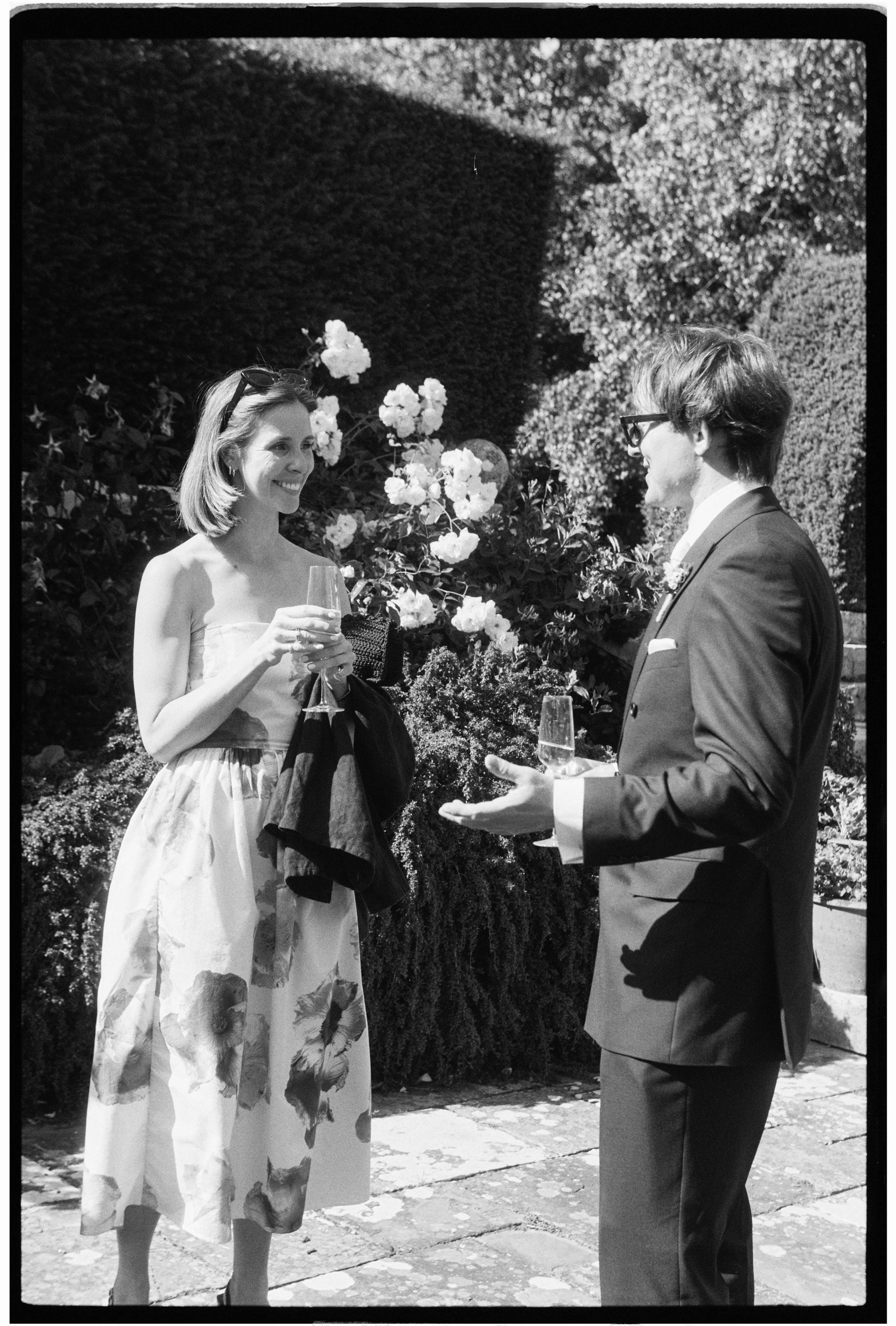 A woman and a man are standing outdoors, dressed in formal attire, holding glasses of wine, and engaging in conversation. The woman is wearing a sleeveless dress with a floral pattern and sunglasses on her head. The man is wearing a suit with glasses