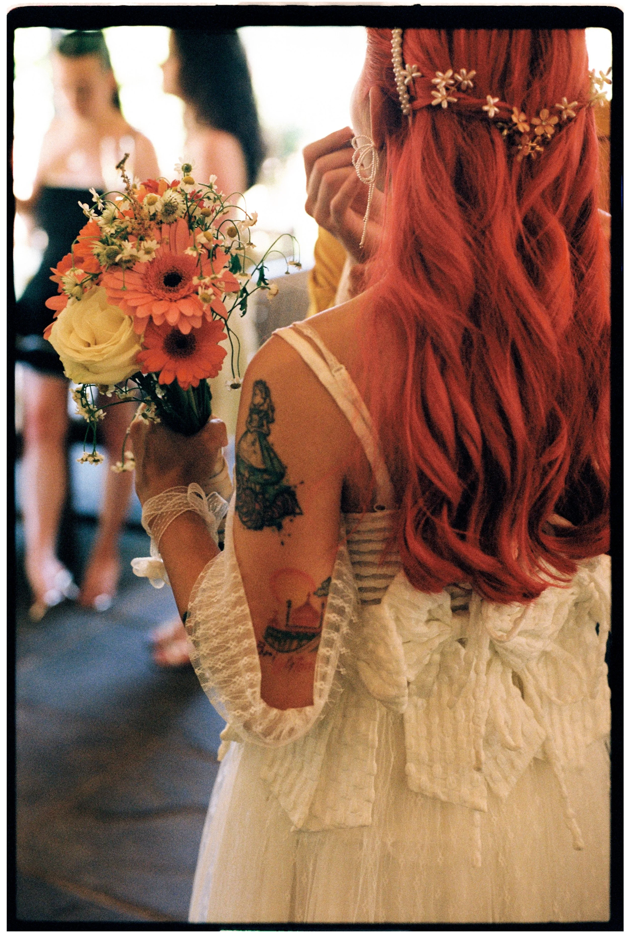 A woman with long red hair holding a bouquet of flowers with orange, white, and yellow blooms at an indoor event, with other women in the background.