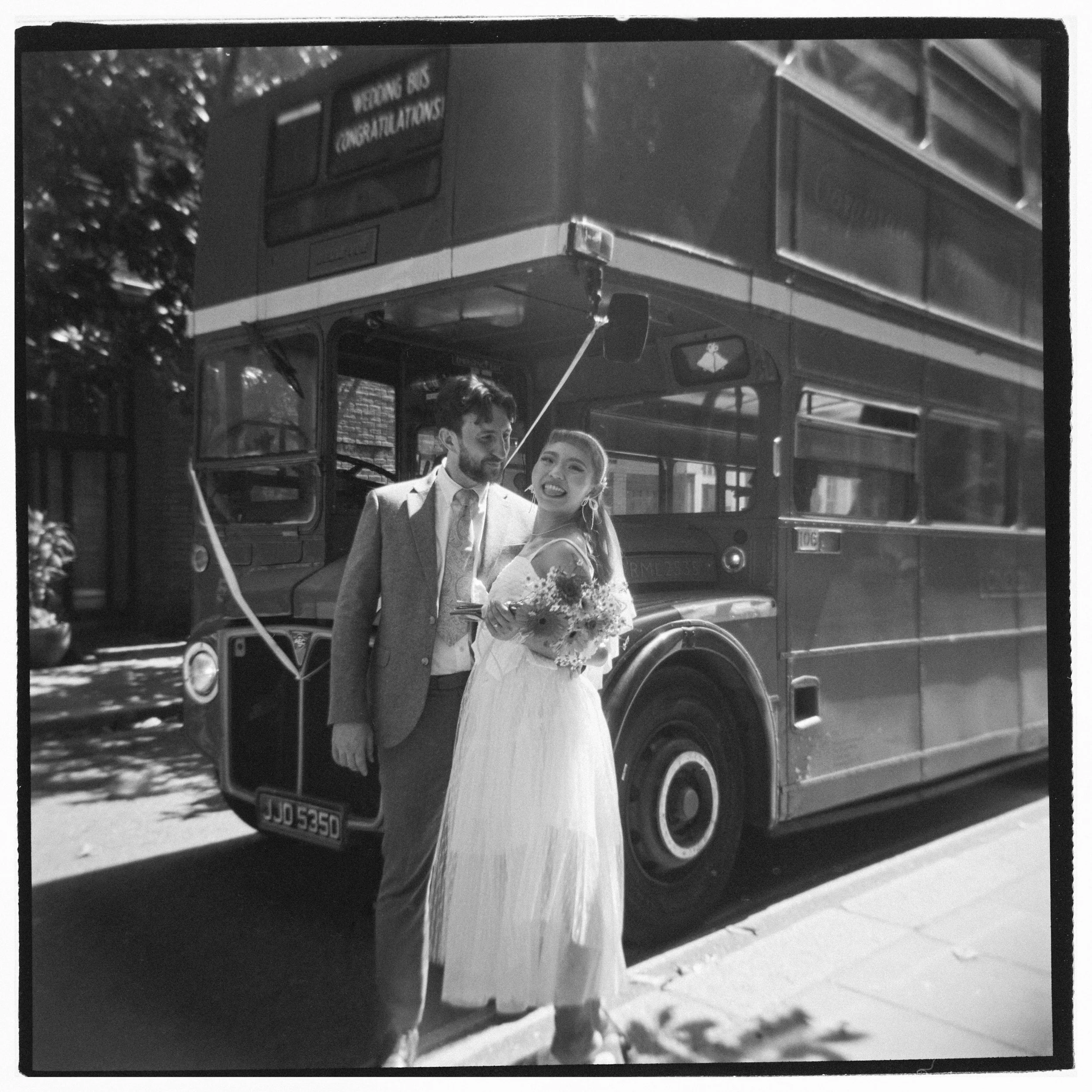 Black and white photo of a double-decker bus with a man and woman smiling in front of it, the woman holding a bouquet, with the bus displaying a message that reads 'Meeting OK! Congratulations'.