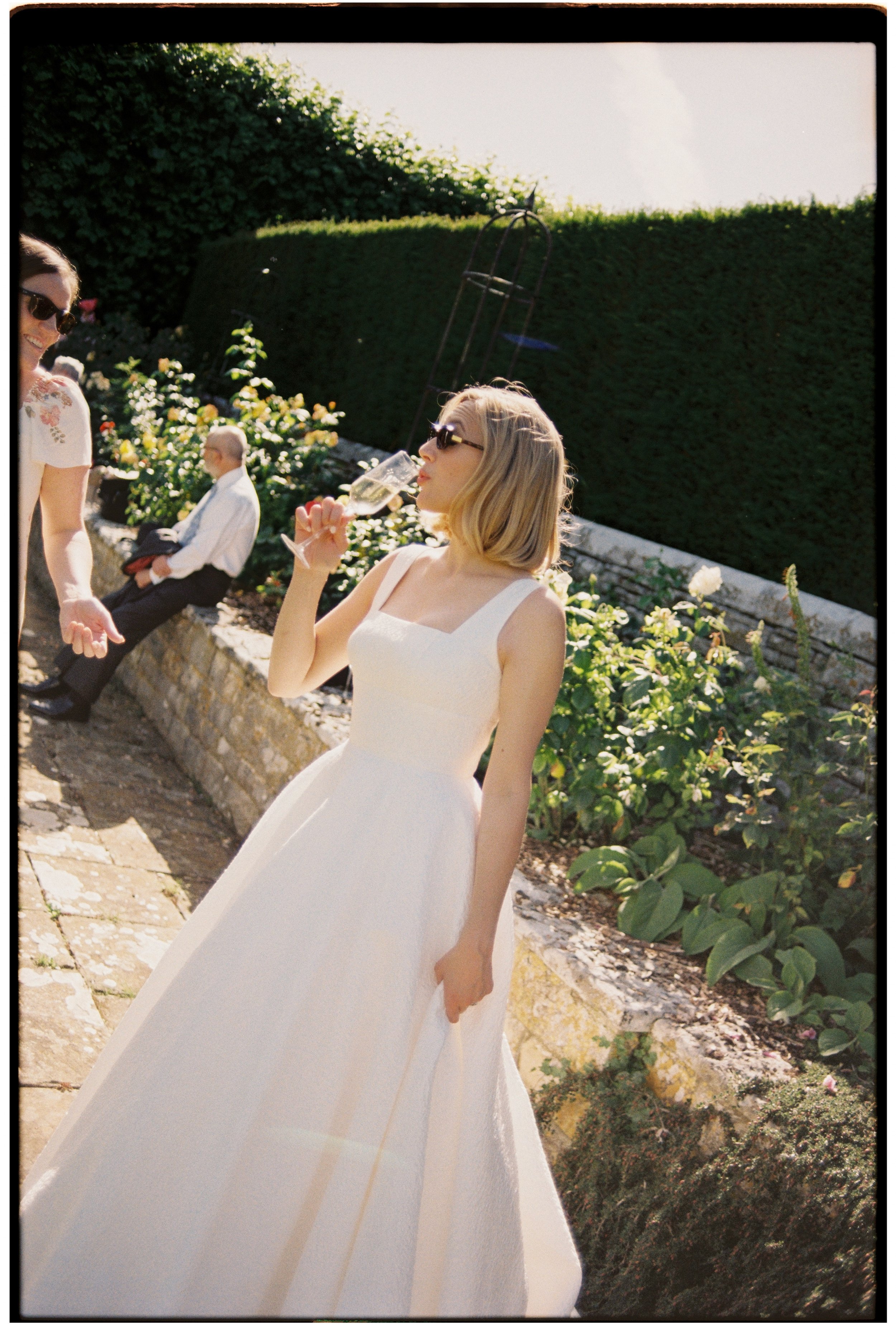 A woman in a white dress and sunglasses holding a champagne glass, standing outdoors in a garden with green plants and flowers, during the daytime.