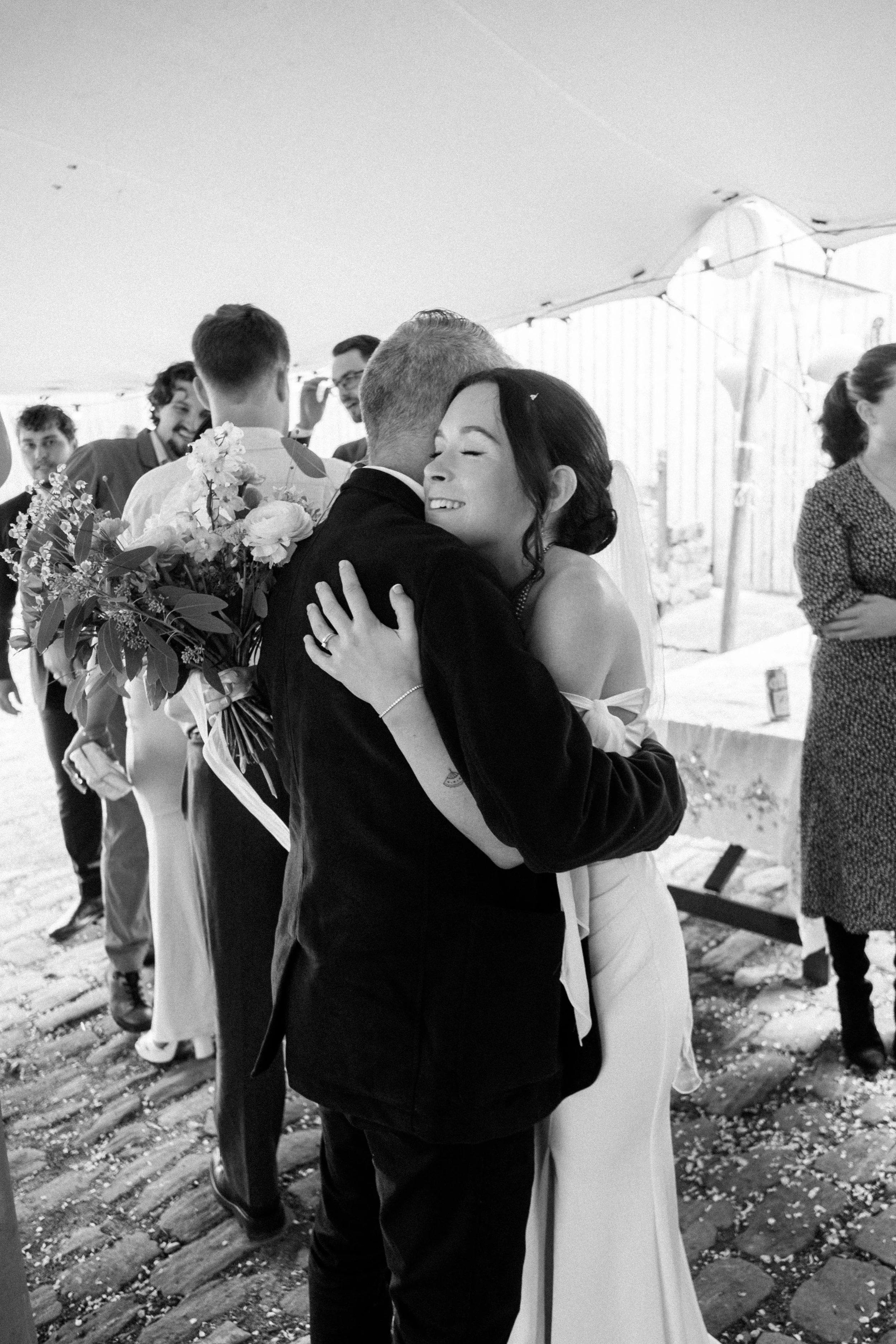 A black and white photo of a wedding reception with a bride and groom hugging, surrounded by friends and family.