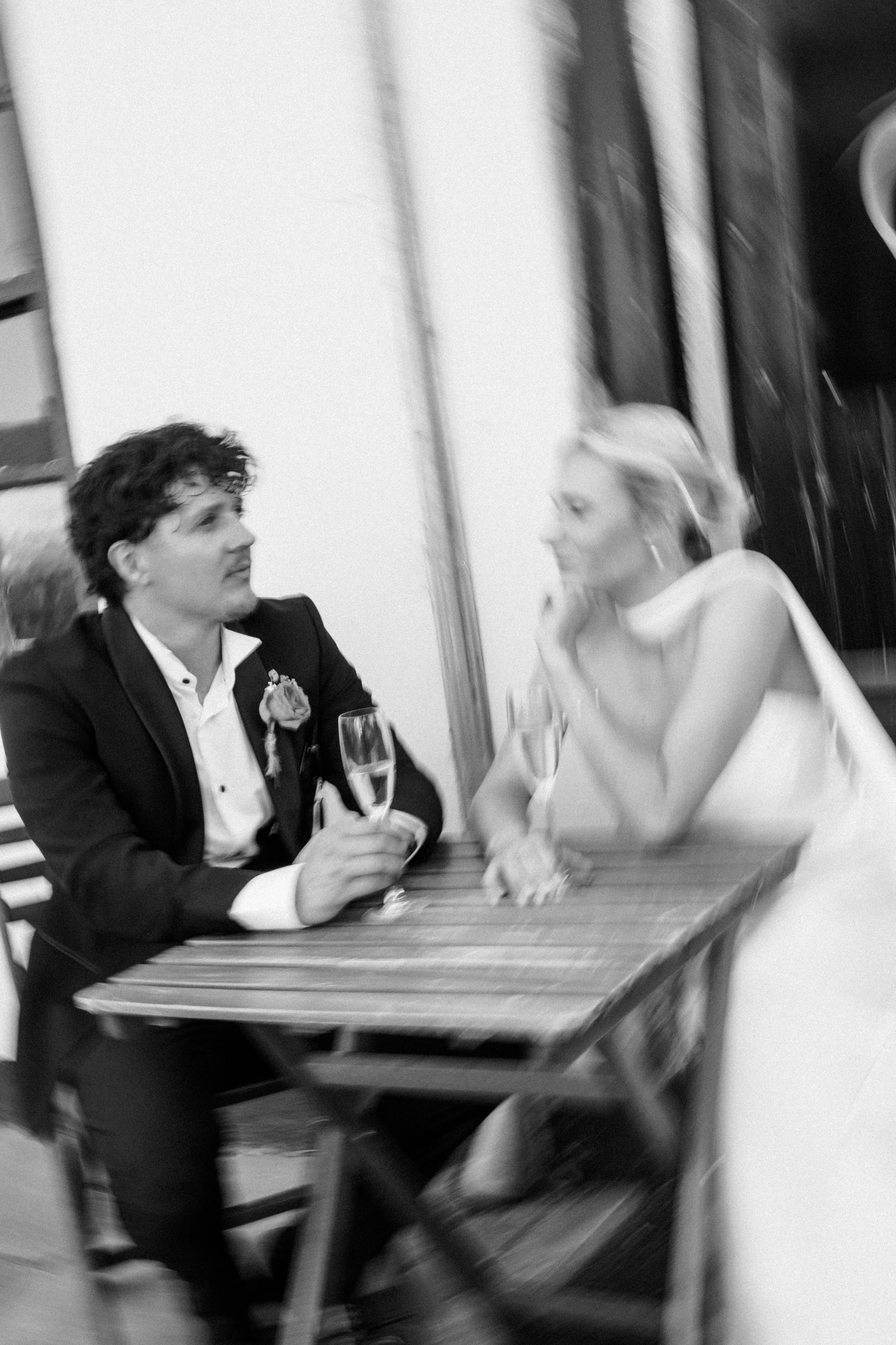 Black and white photograph of a man and a woman sitting at a table, engaged in conversation, with the man holding a glass of champagne. The man is dressed in a suit with a boutonniere, and the woman is wearing a light-colored dress, with her hand resting on her face. The setting appears to be an indoor event or celebration.